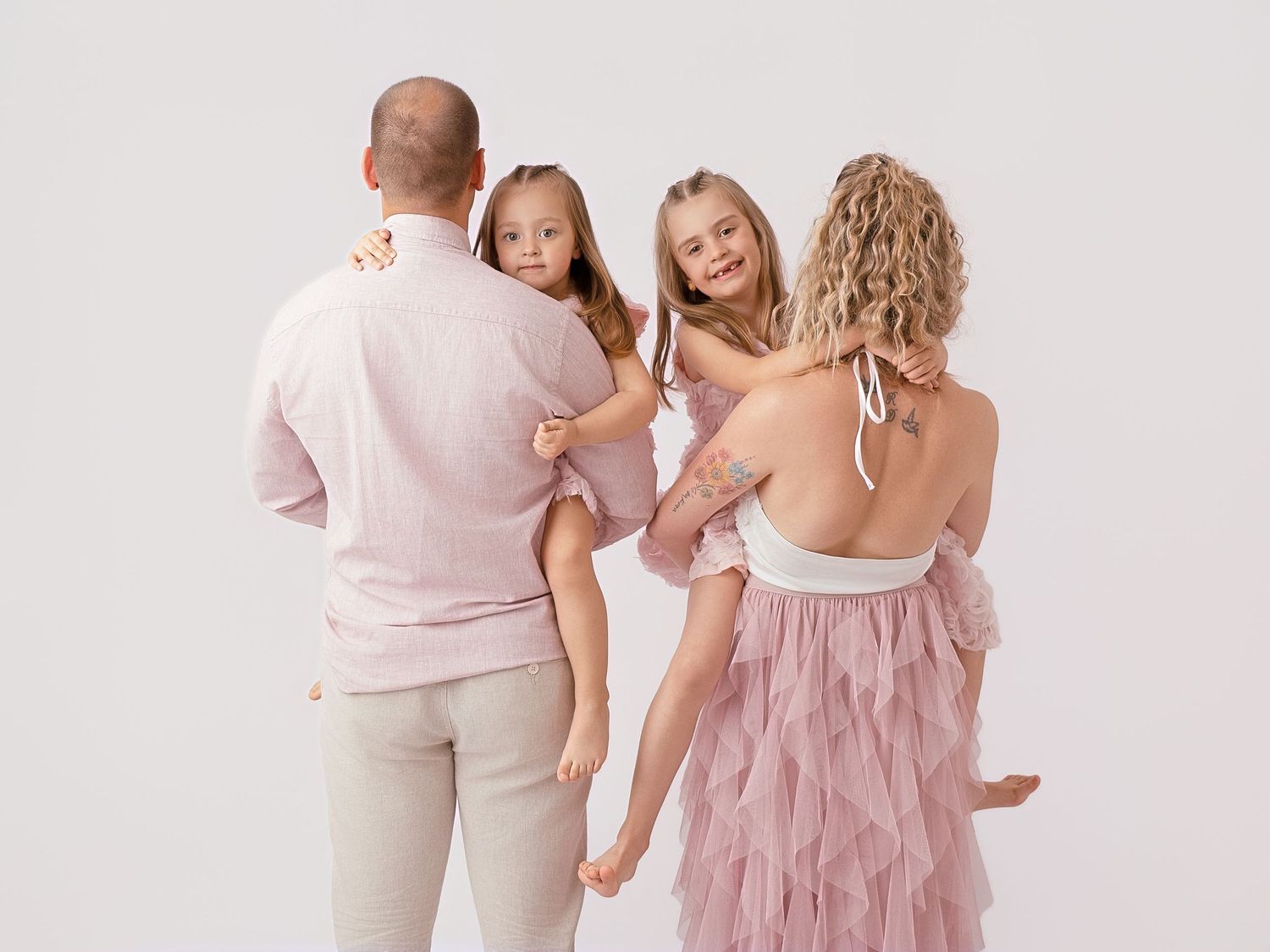 Family of four in pink outfits posing for a portrait, parents holding two young girls against a white background.