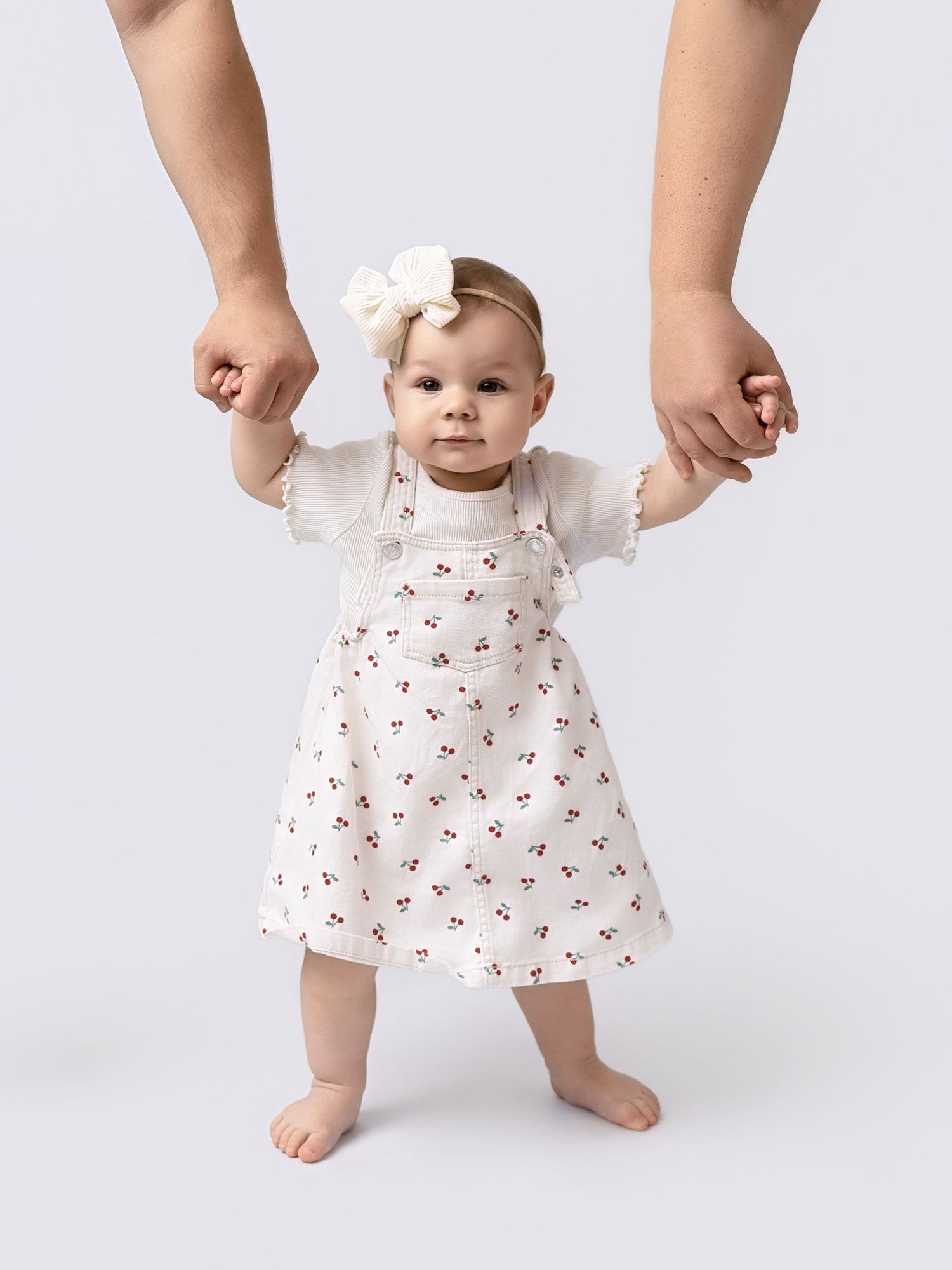 Adorable baby girl in floral cherry dress and white bow headband standing with adult support on both hands.