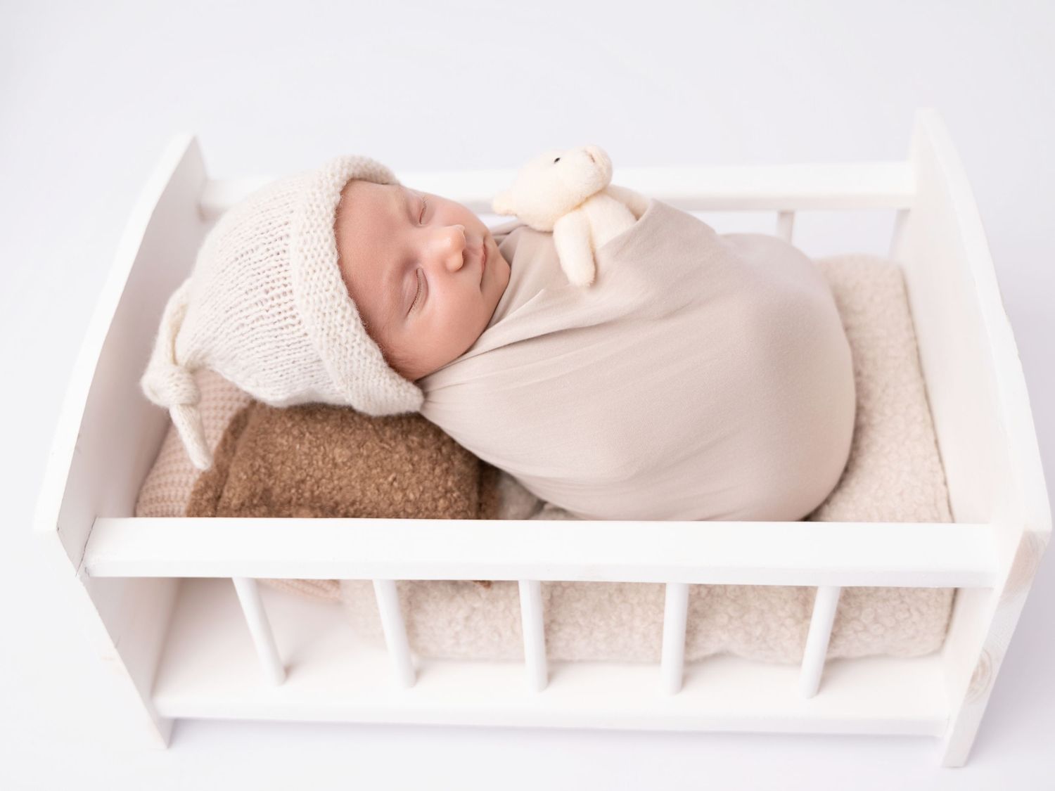 Sleeping newborn baby wrapped in beige swaddle with knit bonnet, holding a tiny teddy bear in a white mini crib.