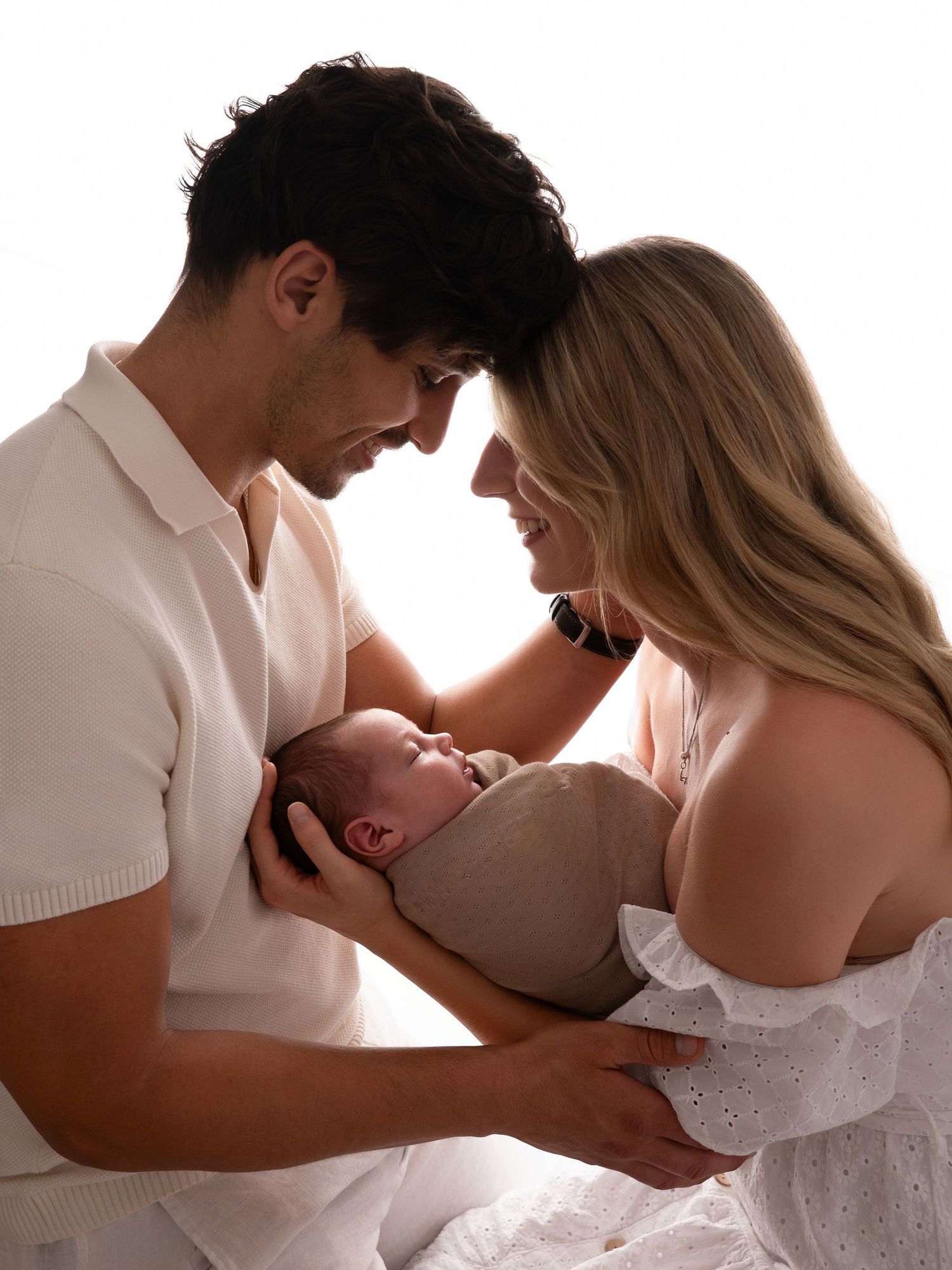 New parents lovingly cradling their sleeping swaddled newborn, foreheads nearly touching in an intimate studio portrait.