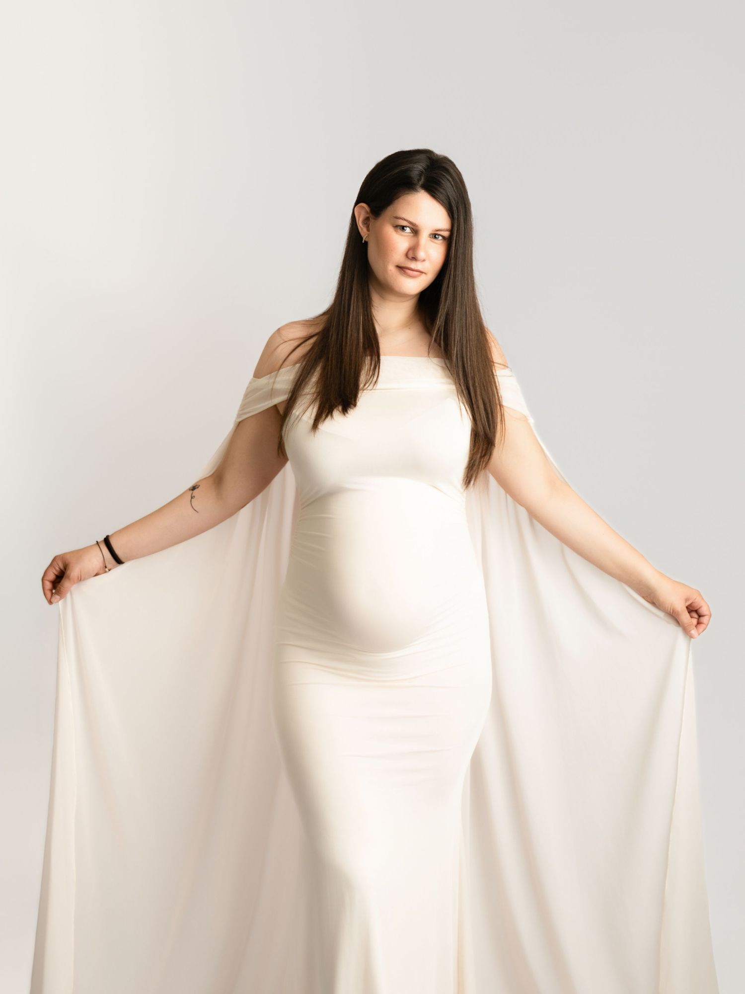 Elegant pregnant woman in white off-shoulder cape maternity gown, studio portrait.