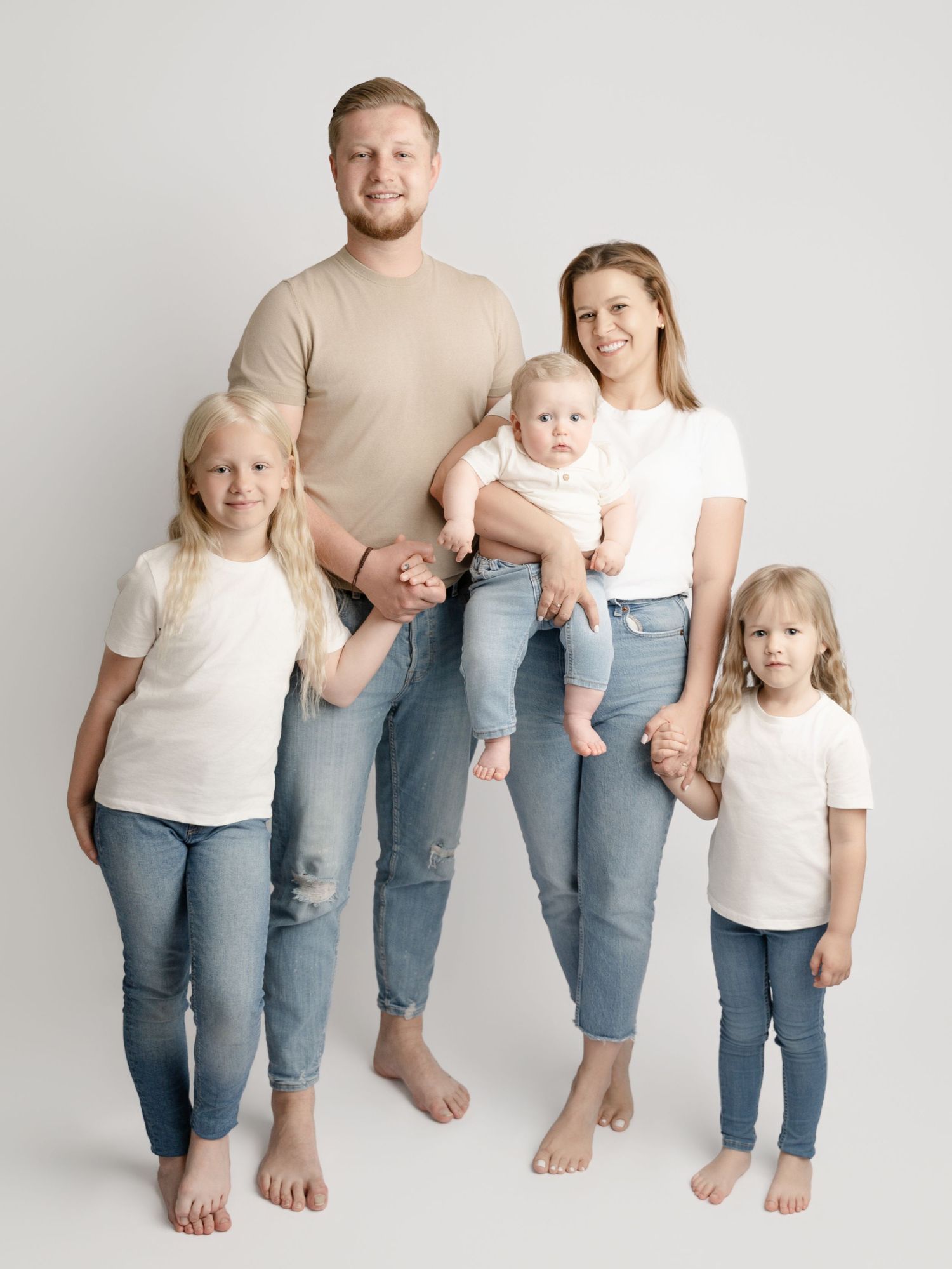 A family of five in coordinating neutral outfits stands together barefoot in a clean studio setting.