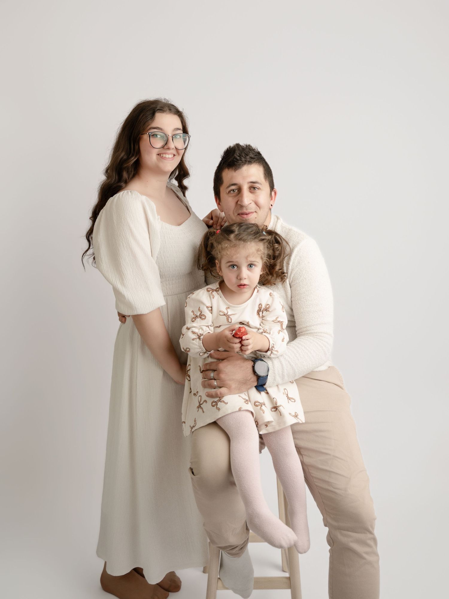 Family of three in cream outfits posing with young daughter on stool against white studio background.