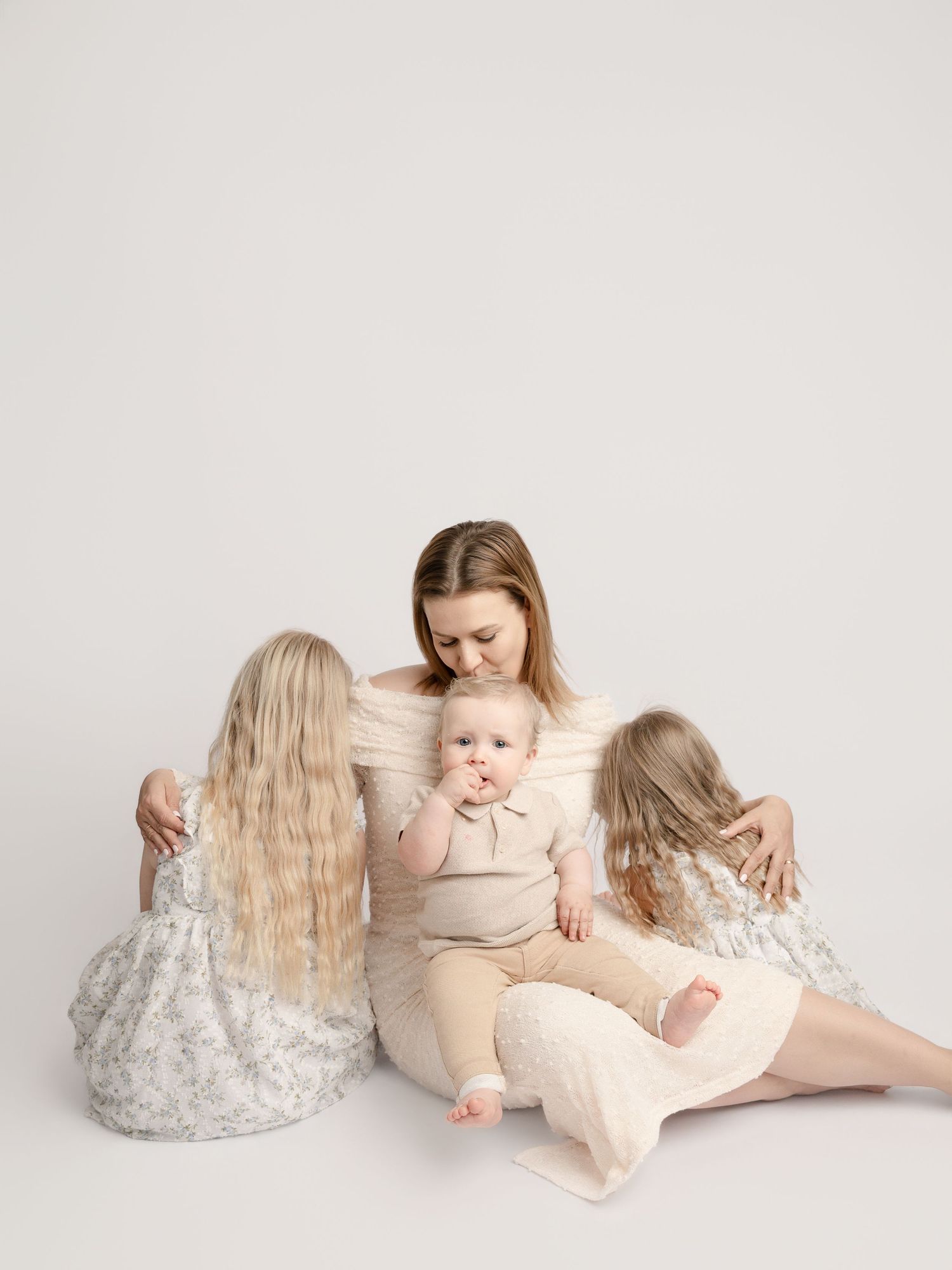 Mother sitting on floor with baby and two daughters in soft neutral tones studio family portrait.