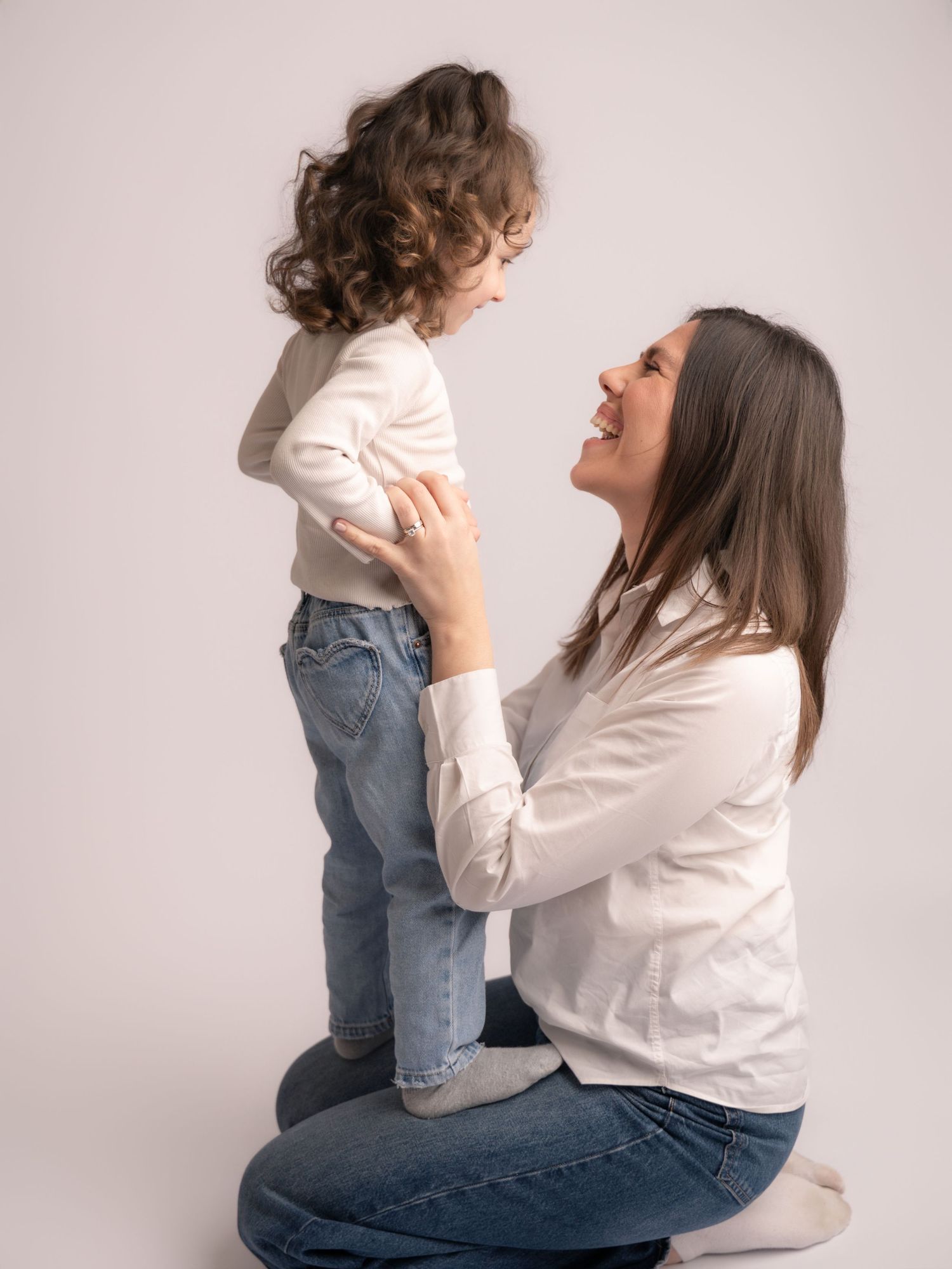 Laughing mother kneels holding her curly-haired toddler in white top and jeans in studio.