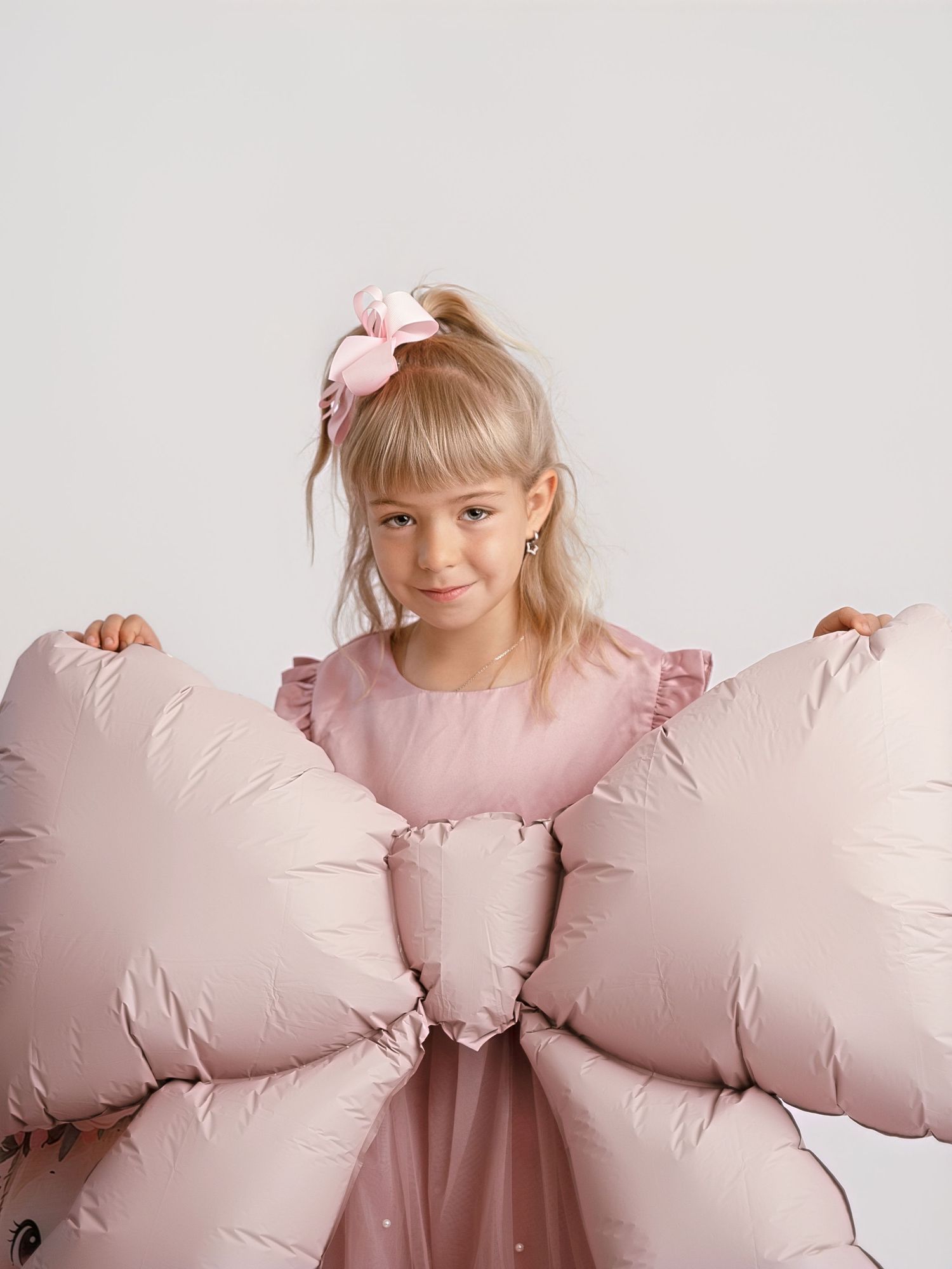 Young blonde girl in pink dress holding large pink bow-shaped pillow with matching hair accessory.