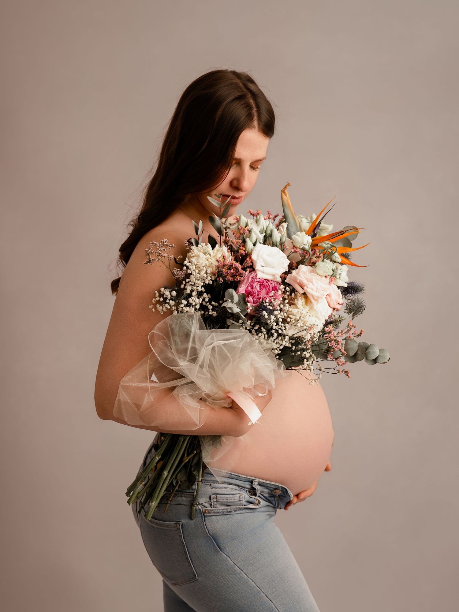 Pregnant woman in jeans holding a colorful floral bouquet, showing her baby bump in a studio maternity photo shoot.