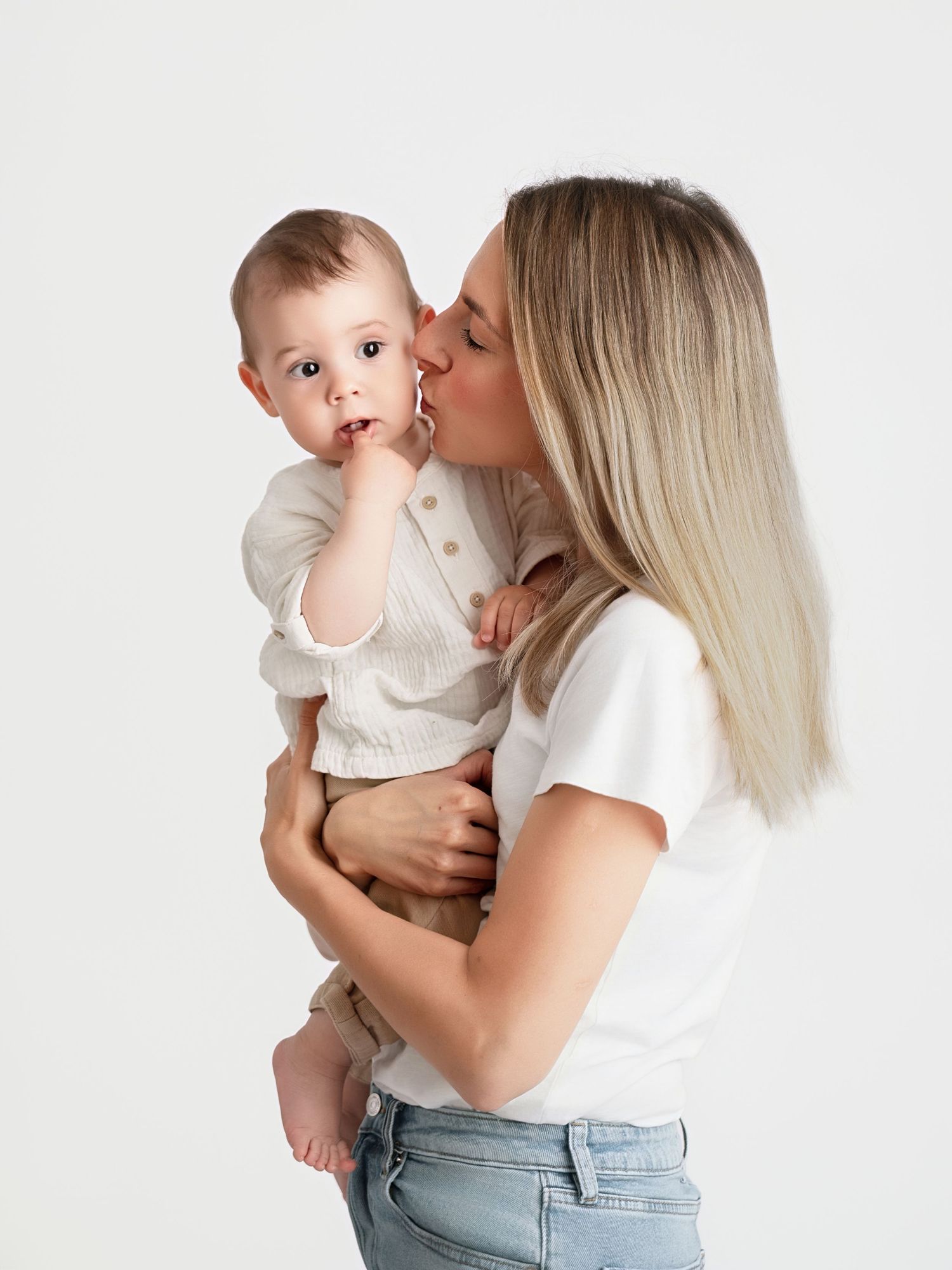 A blonde mother in a white tee and jeans kisses her baby boy on the cheek against a white background.