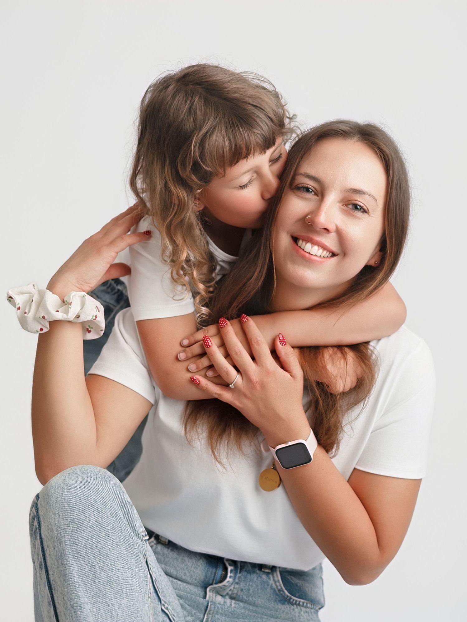 A young girl hugs and kisses her smiling mother from behind, both wearing white t-shirts against a light background.