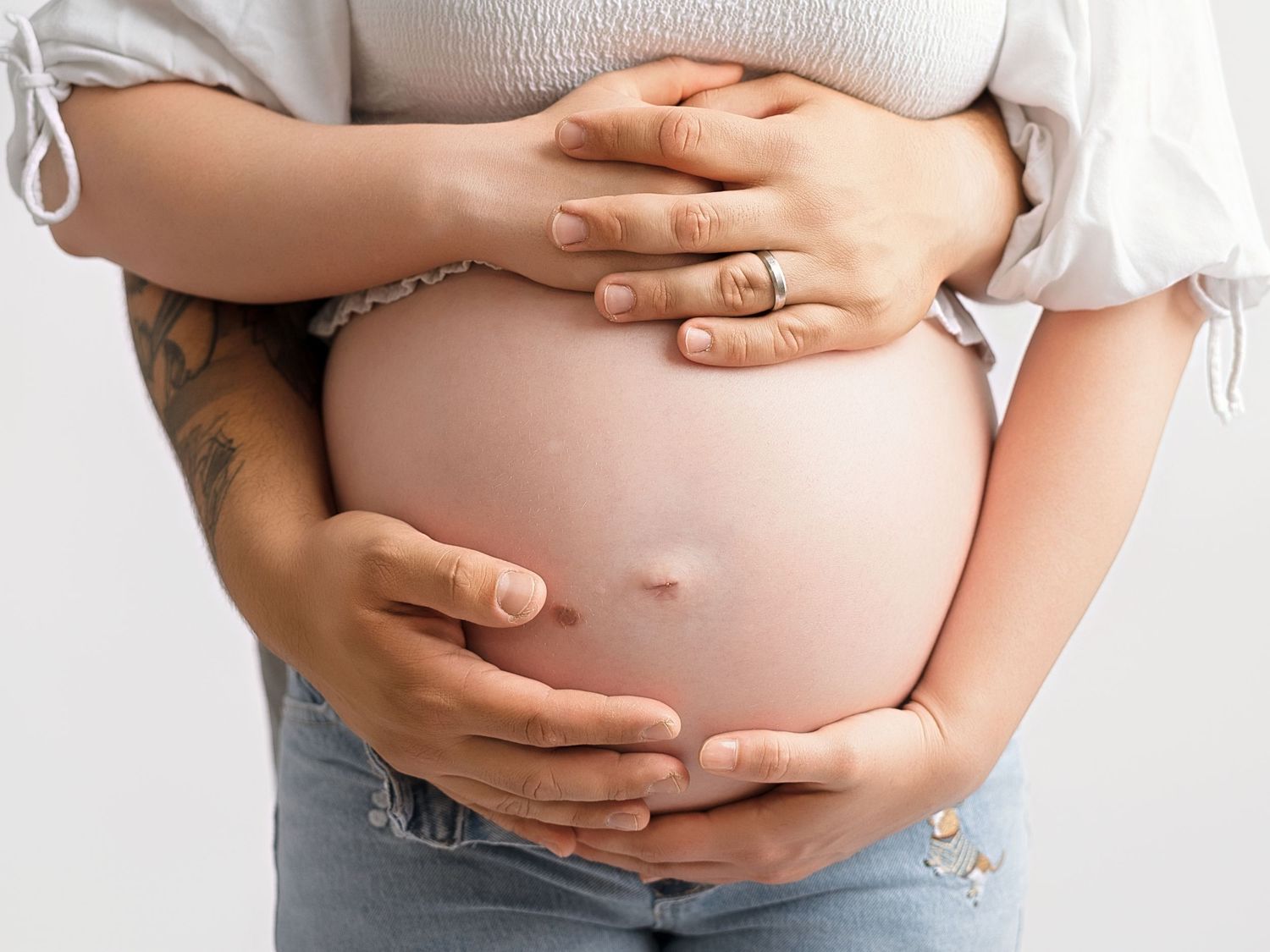 Pregnant woman in jeans cradling her bare baby bump with both hands, showing a tattoo on her arm.
