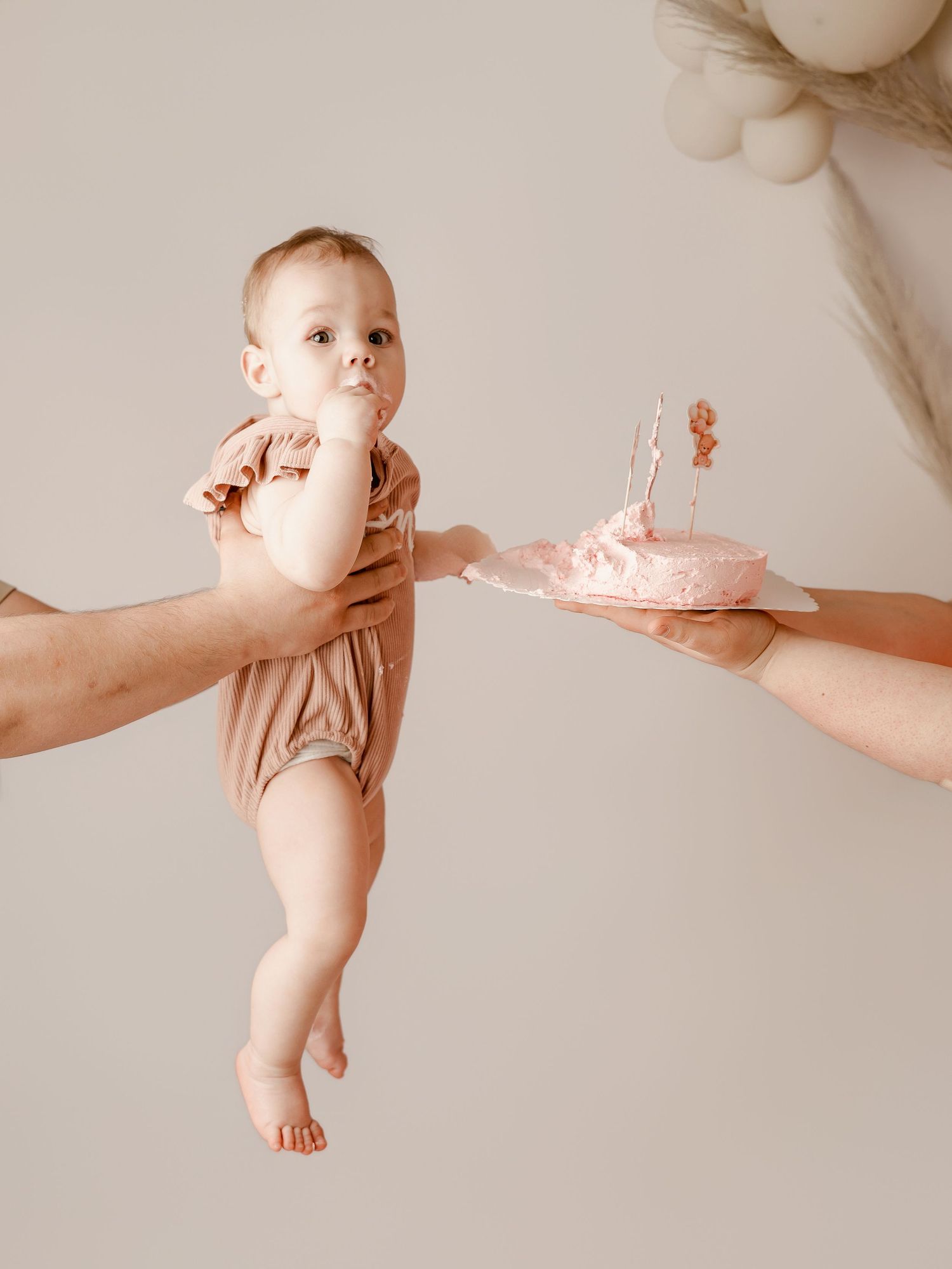 Baby in pink romper held by adult hands next to a small pink smash cake with candle for first birthday.