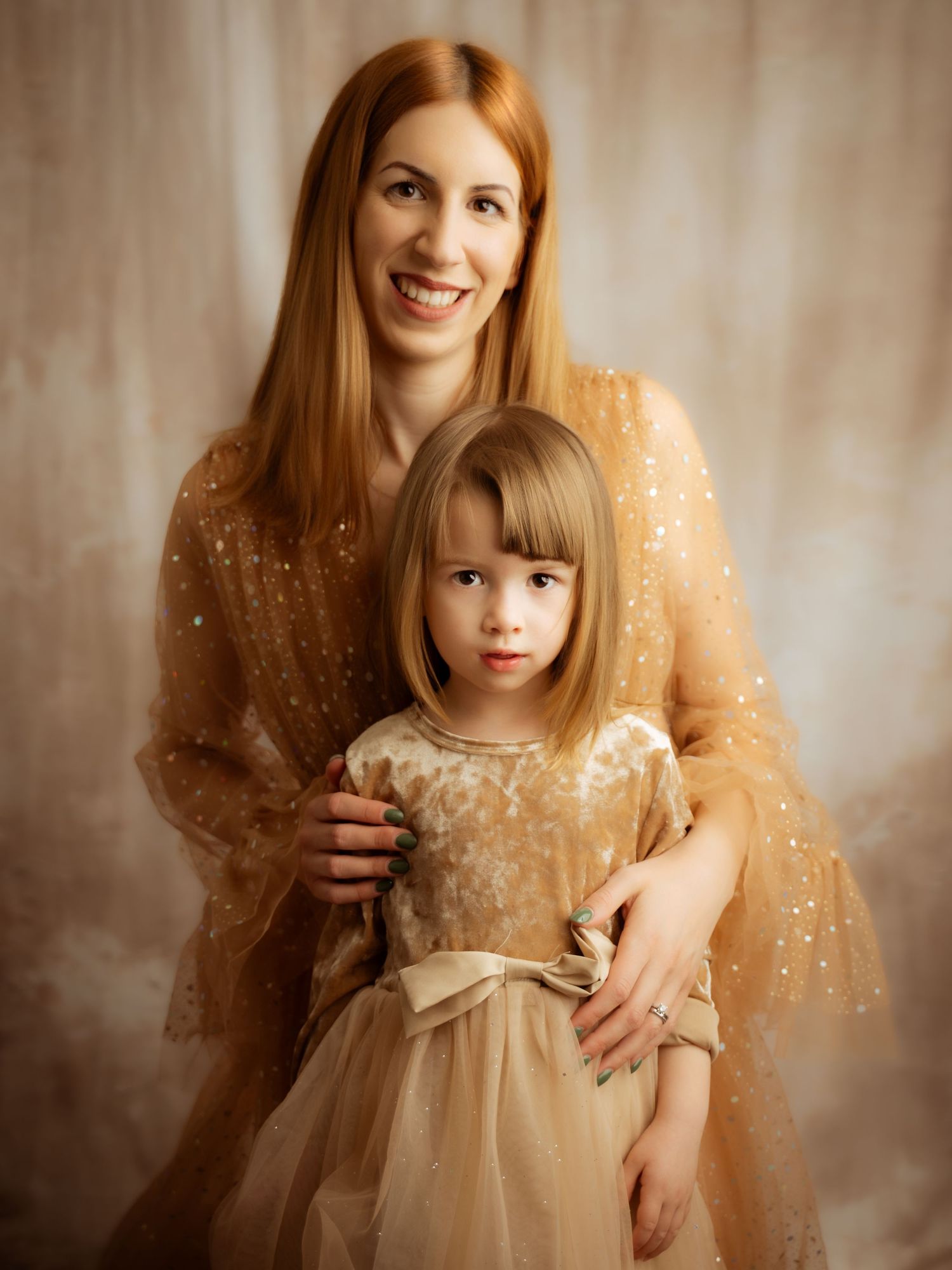 A red-haired mother embraces her young daughter, both wearing elegant gold sparkle dresses in a studio.