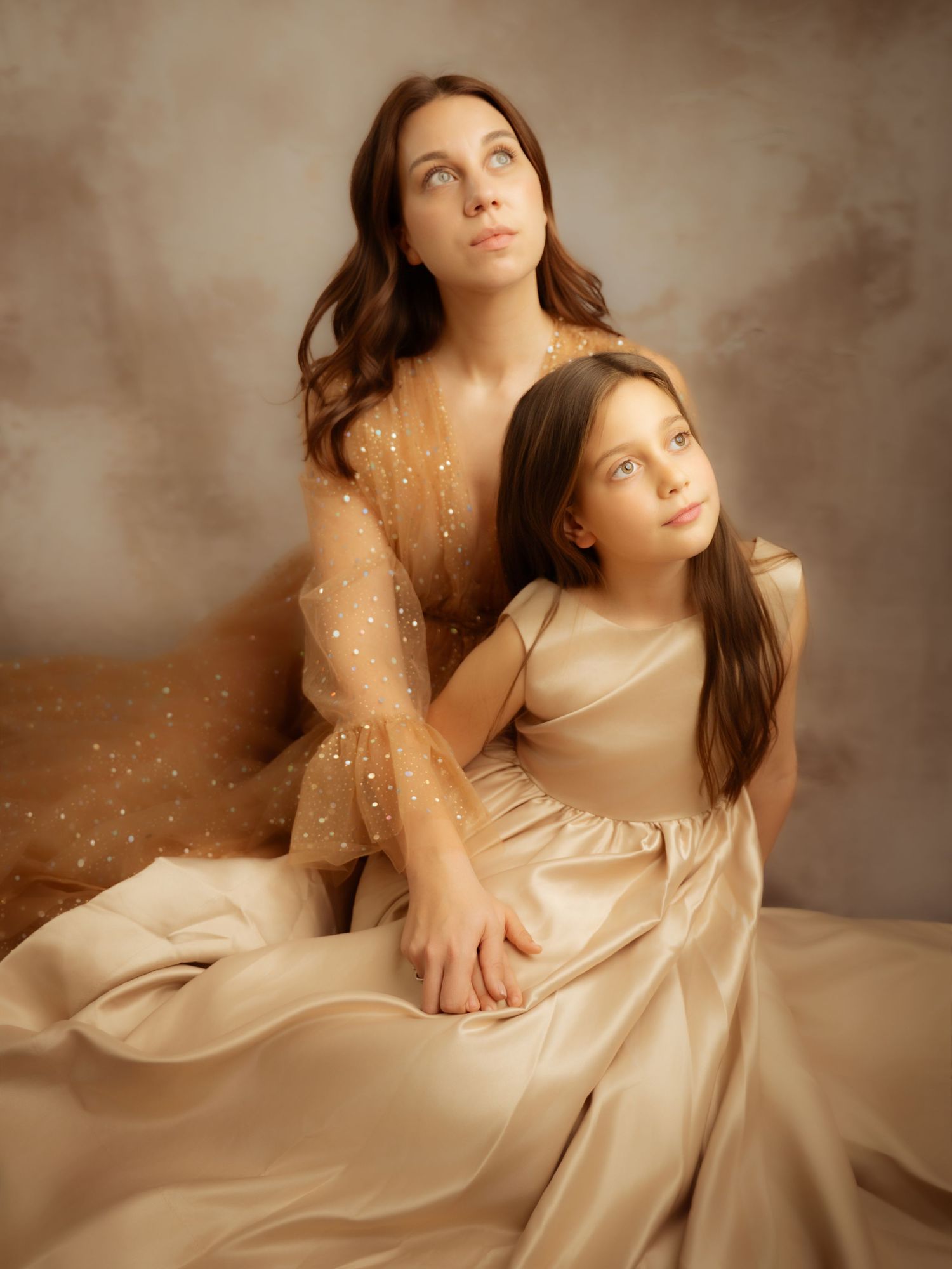 Mother and daughter in elegant golden beige dresses posing together in a classic fine art portrait studio photo.