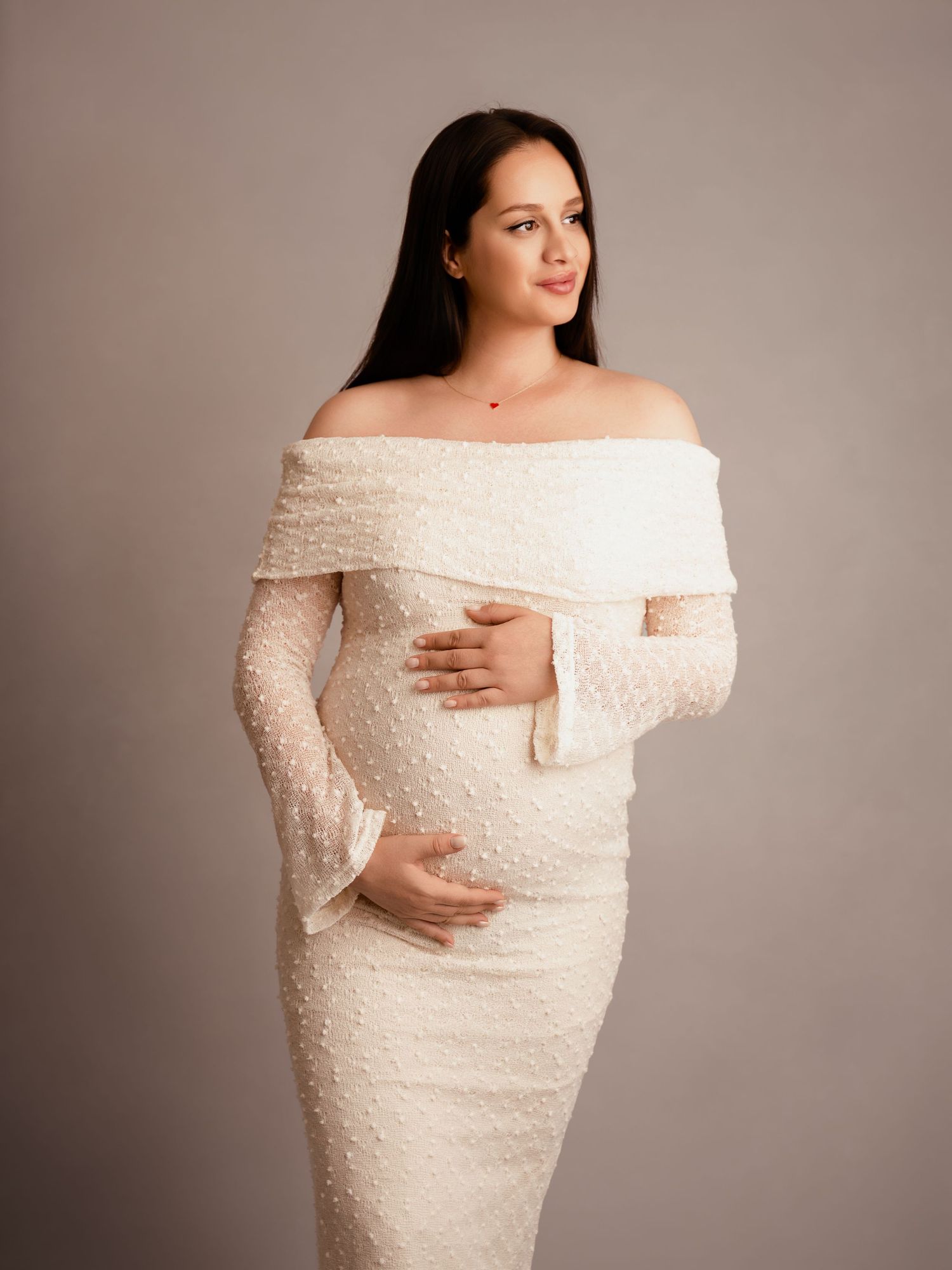 Pregnant woman in elegant white off-shoulder lace maternity gown cradling baby bump in professional studio photo.