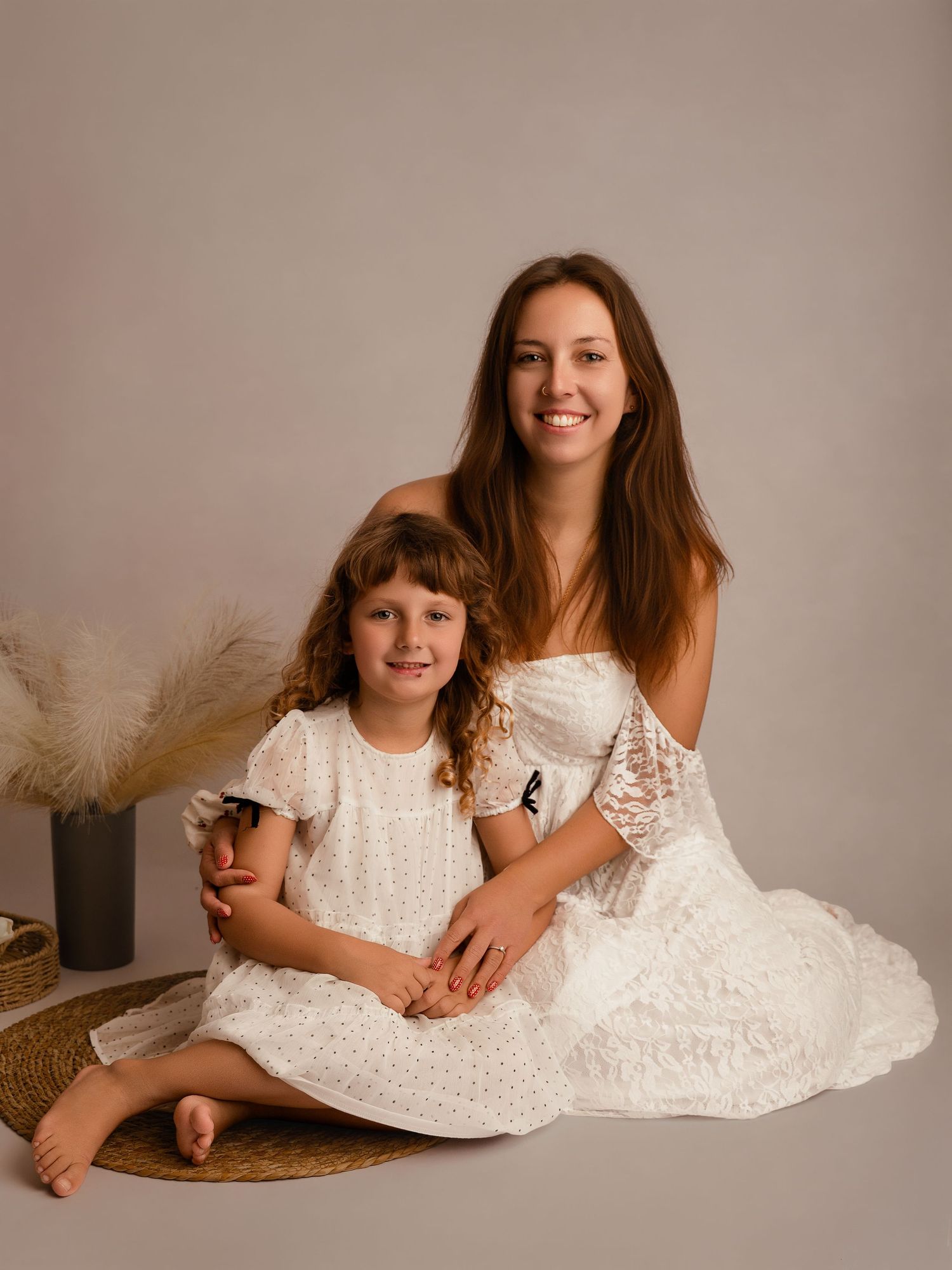 Woman and young girl in matching white lace dresses pose together for a studio portrait photo.
