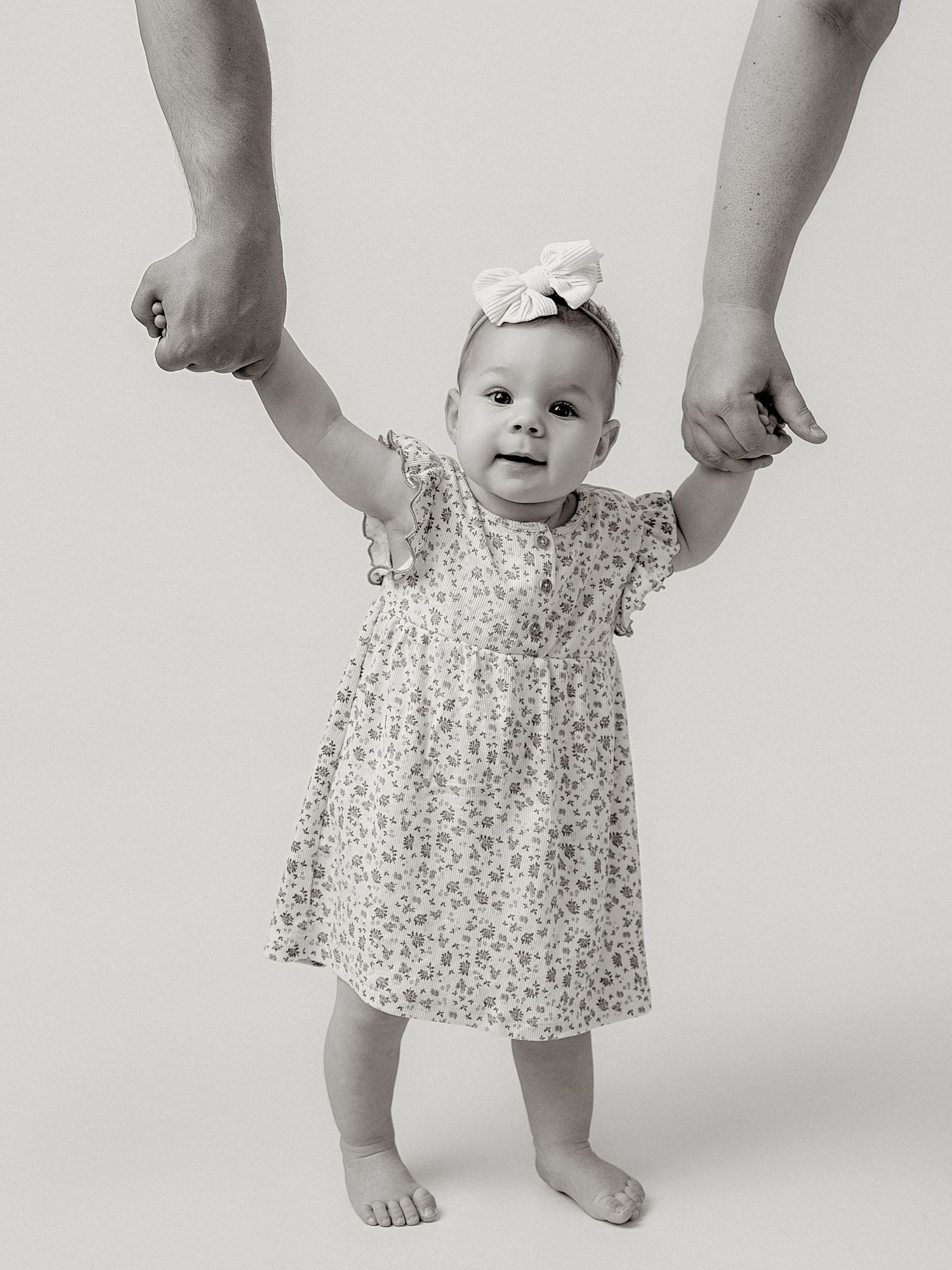 Black and white photo of a baby girl in a floral dress and bow headband, learning to walk while holding adult hands.