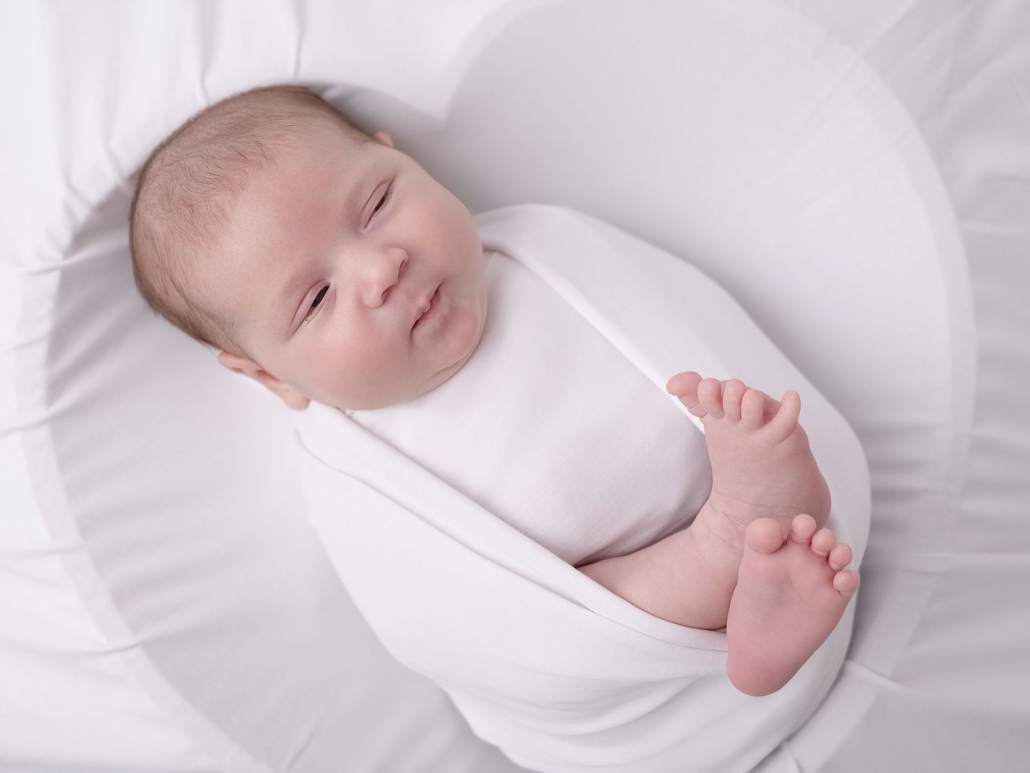 Peaceful newborn baby swaddled in white wrap, smiling while sleeping on a white pillow with one foot peeking out.