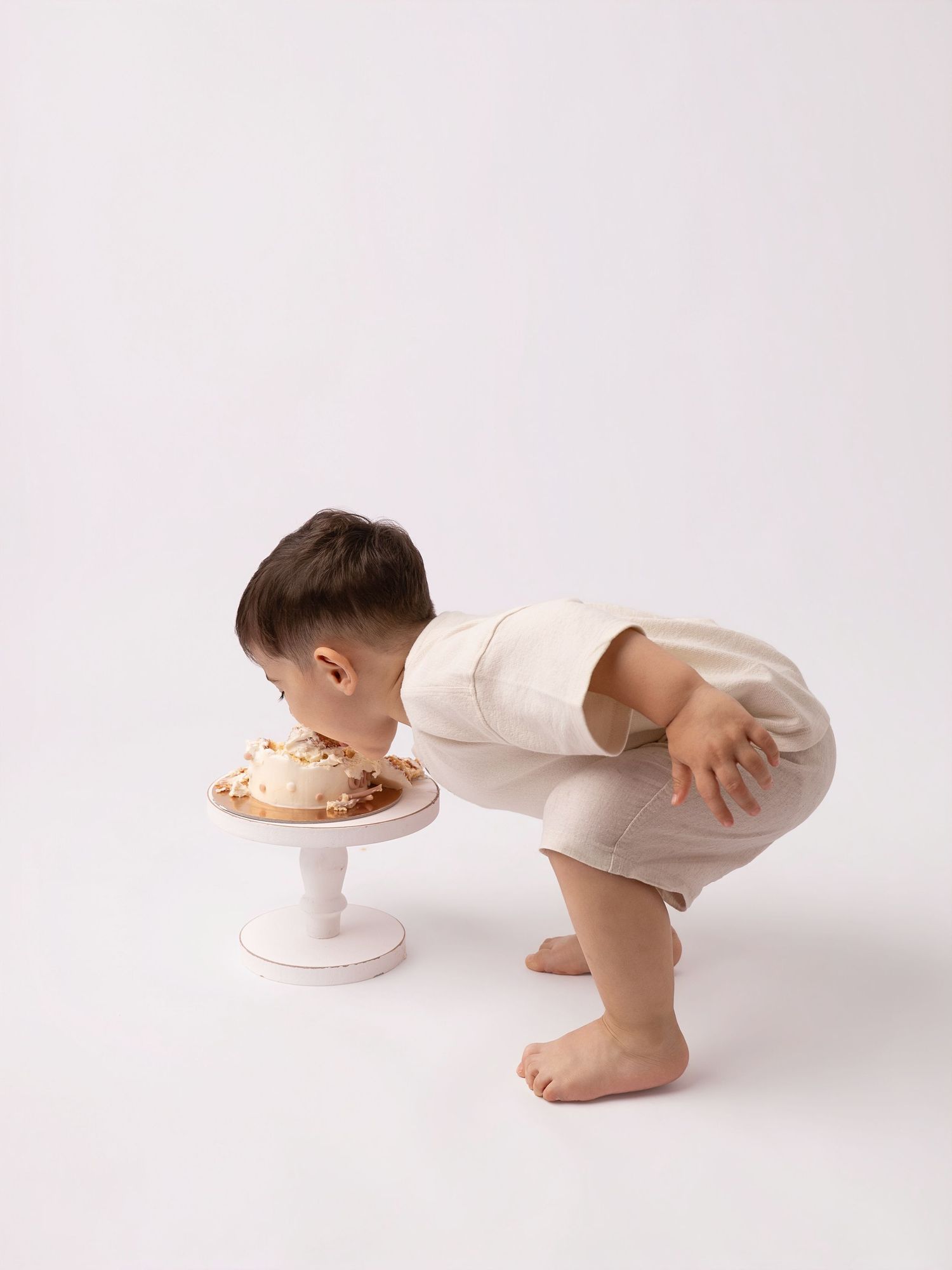 Toddler boy in cream outfit leaning over a white cake smash cake on a pedestal stand against white background.