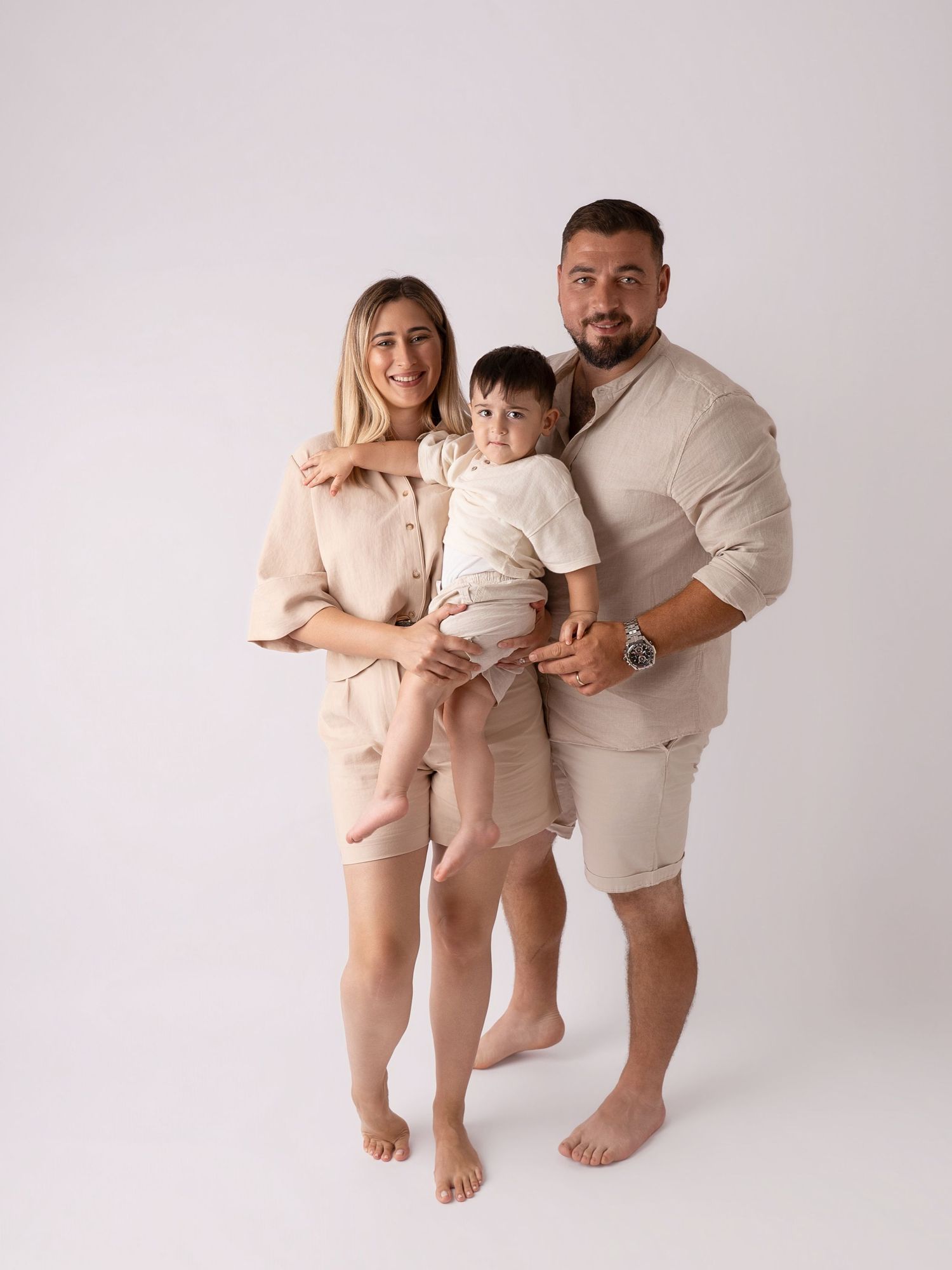 Happy family of three in coordinating beige outfits standing together in white studio, holding young toddler.