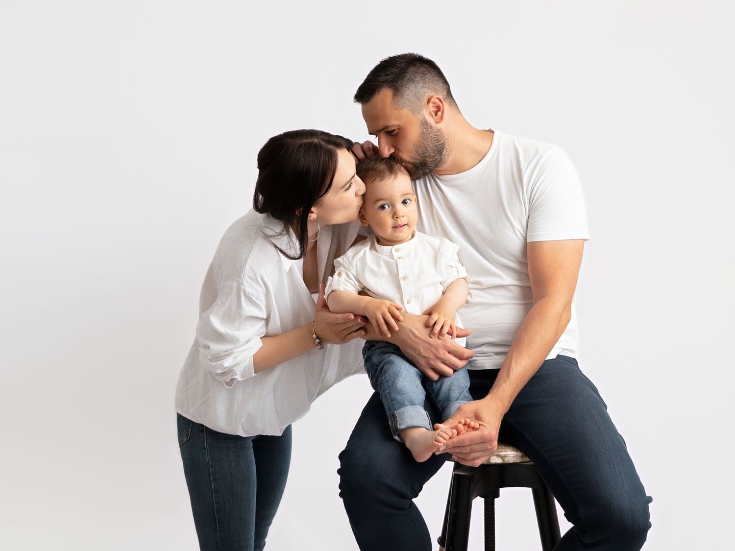 Parents in white shirts kissing their baby on a stool against a white studio background.