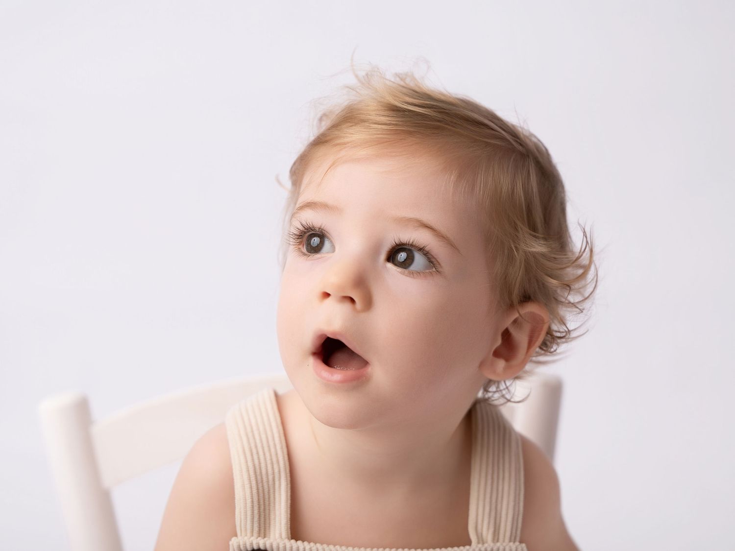 Adorable blonde toddler with wide eyes and open mouth sitting in a white chair against a light background.