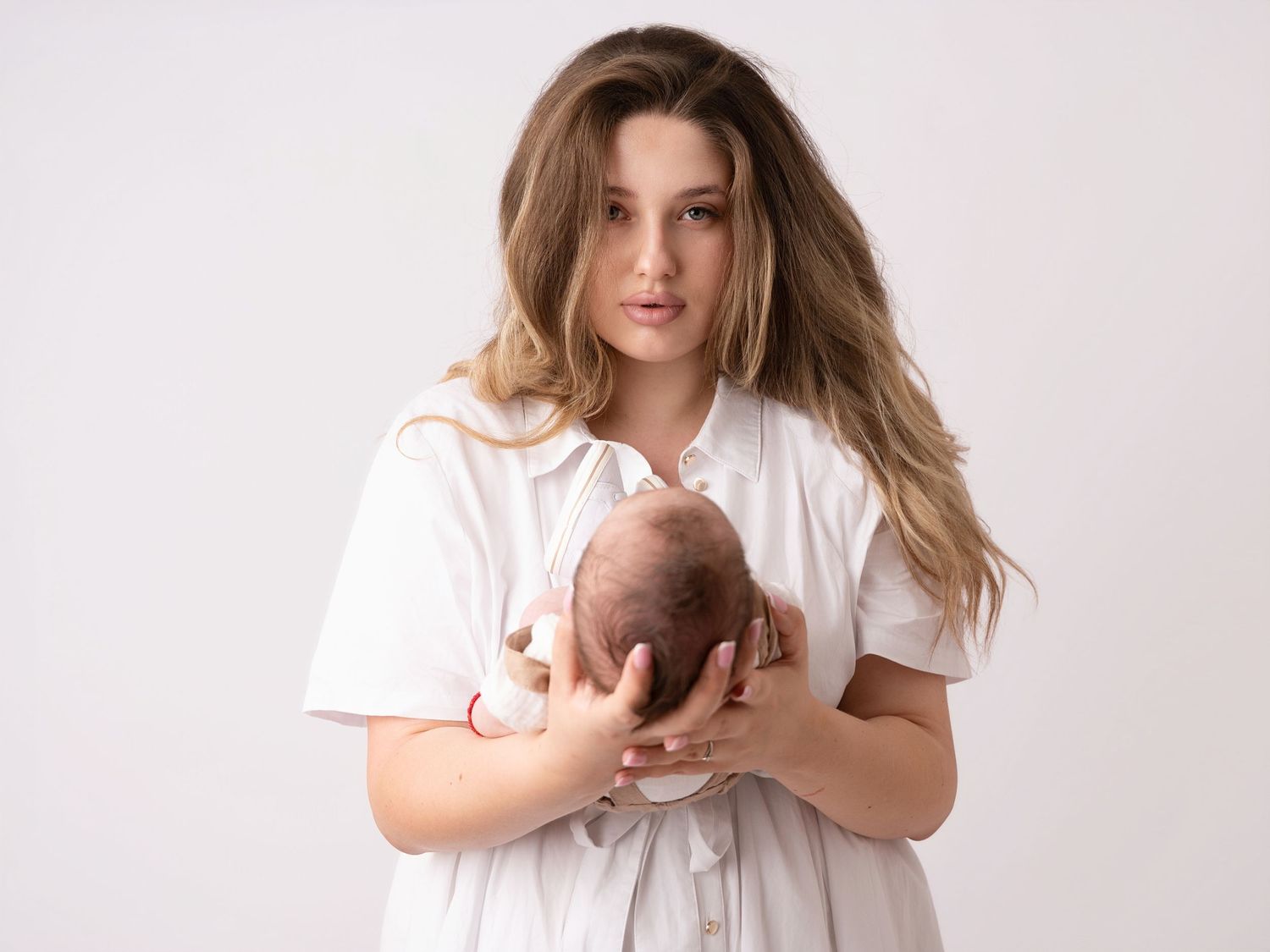 Young woman in white dress tenderly holds newborn baby against white studio background.