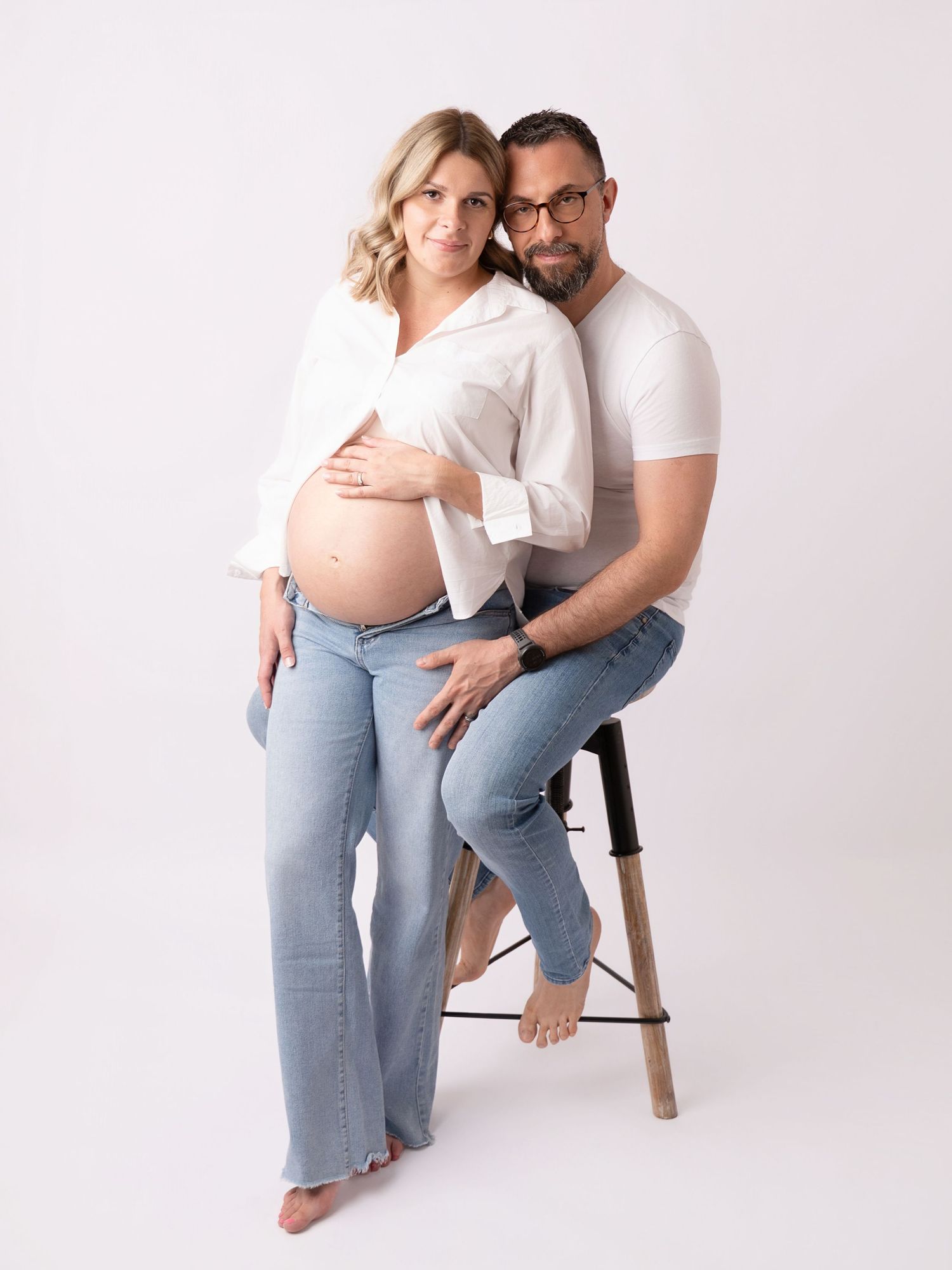 Couple maternity photo shoot, pregnant woman in white shirt and jeans with partner on stool, white studio background.