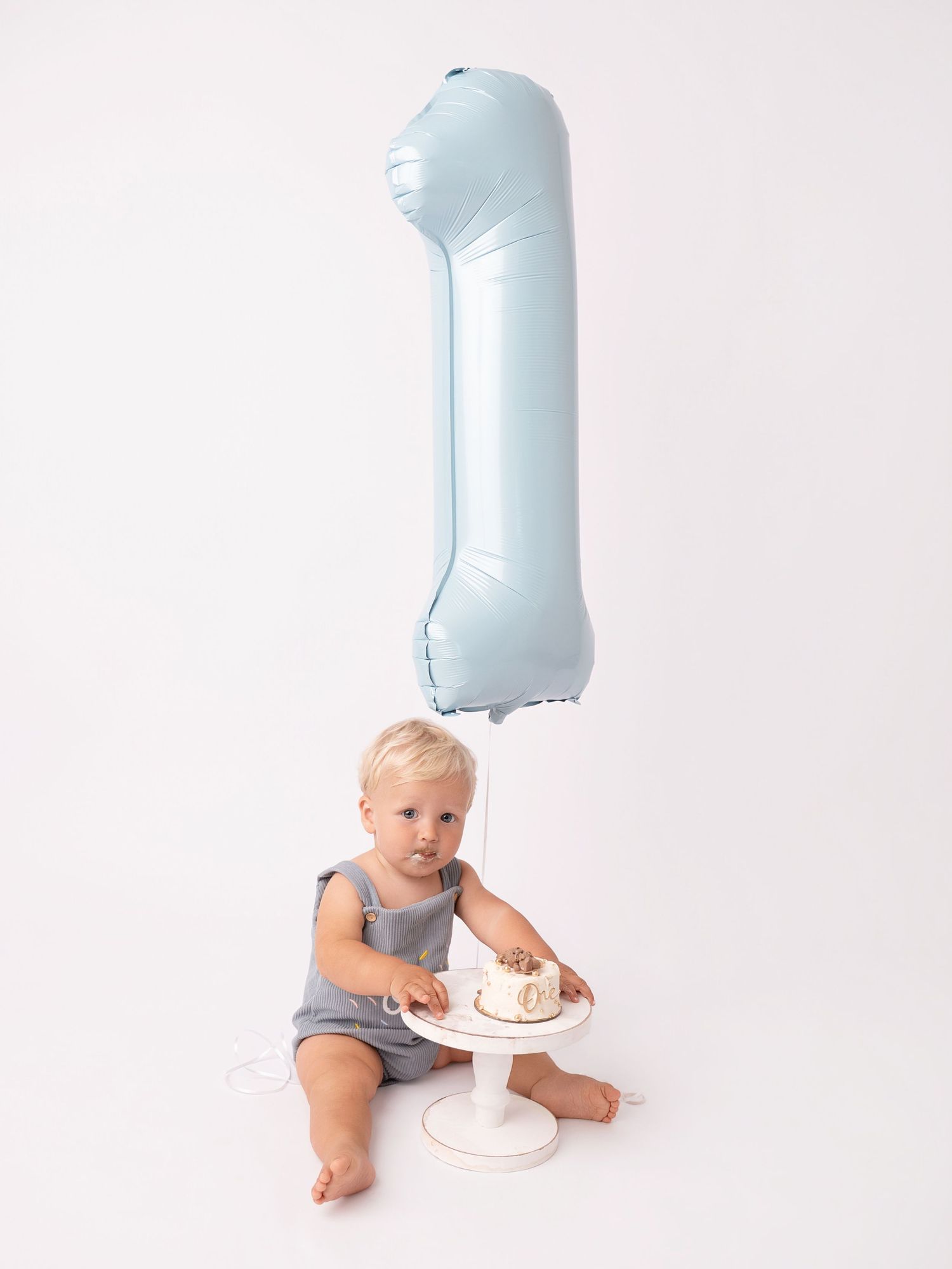Baby boy in gray romper sitting on floor with smash cake and blue number 1 balloon for first birthday.
