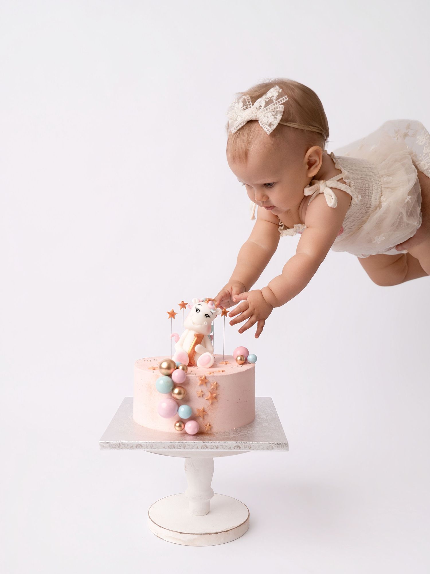 Curious baby girl in white dress reaching into pink first birthday cake on white pedestal stand.