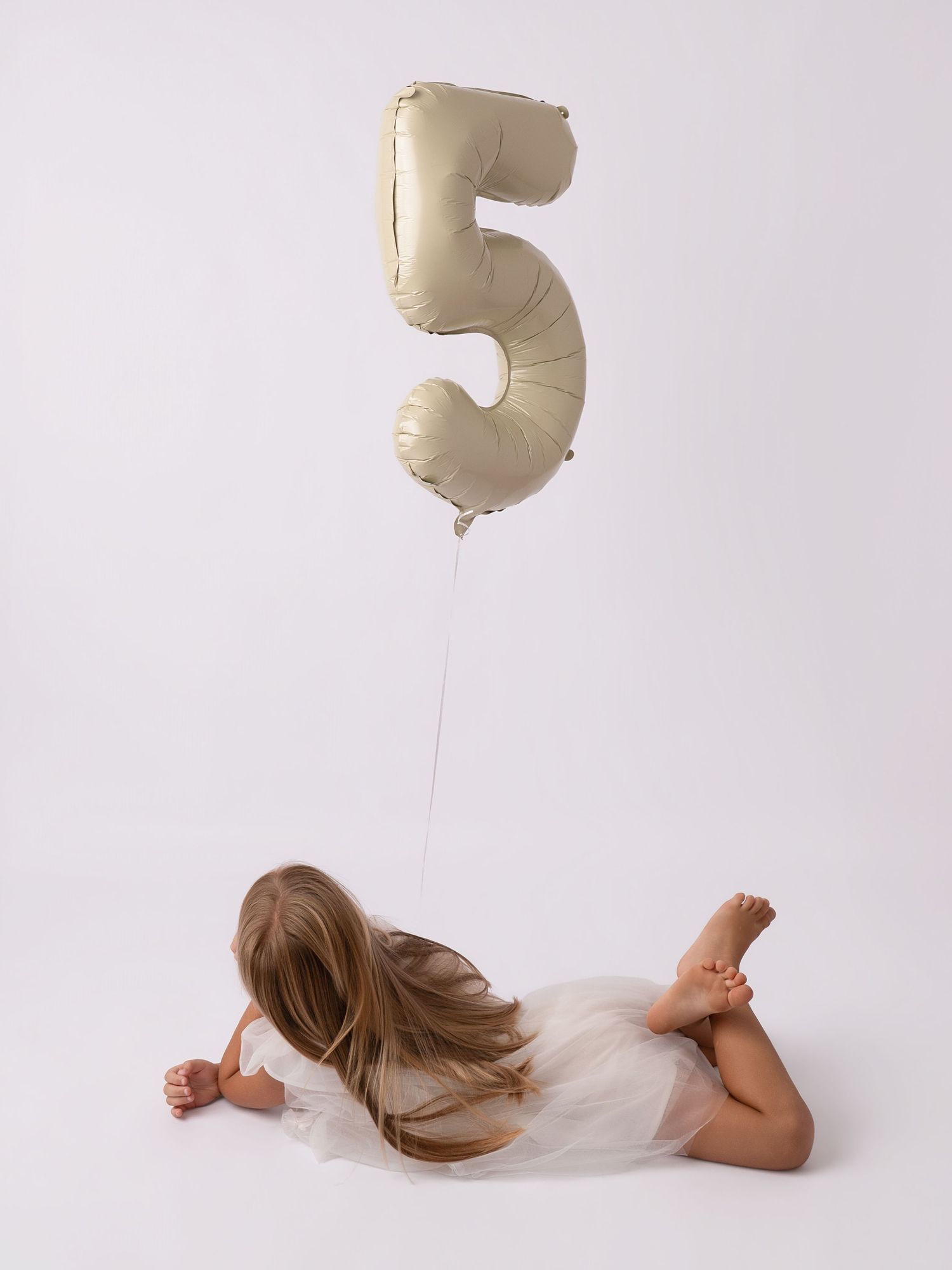 A young girl lying down holding a gold number 5 balloon, wearing a white tutu dress for her 5th birthday celebration.