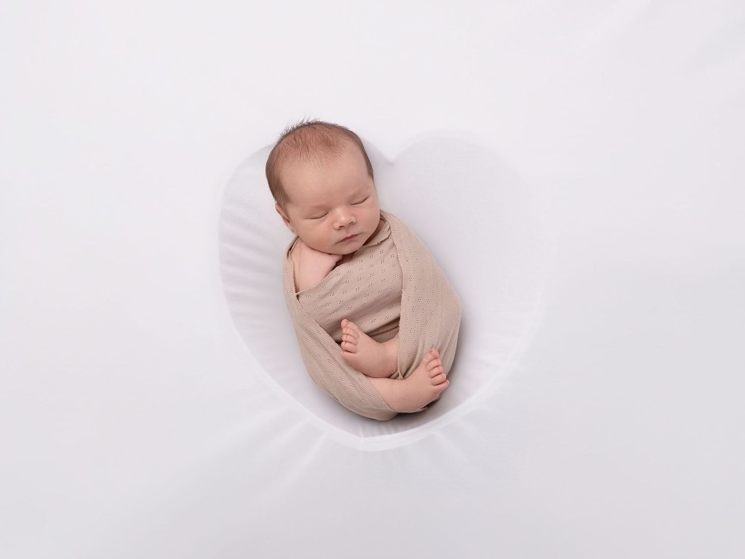 Newborn baby swaddled in beige wrap sleeping peacefully inside a white bowl prop during professional newborn photography session.