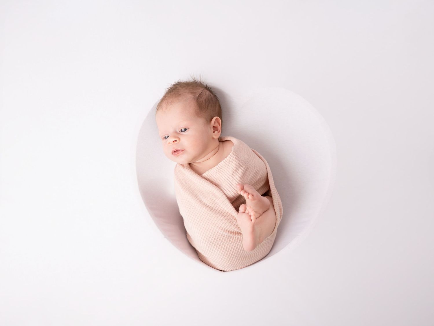 Newborn baby swaddled in pink wrap lying in a round white bowl prop for a studio photography session.