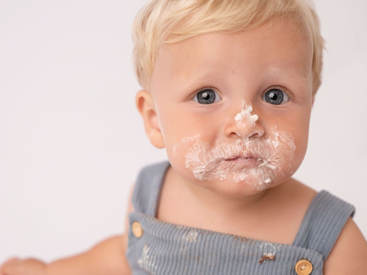 Adorable blonde toddler with cake smeared on face wearing gray overalls during first birthday smash cake photo session.