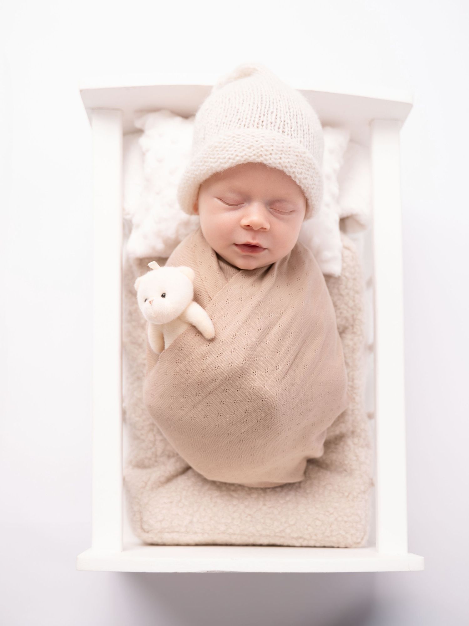 Newborn baby in a beige swaddle and knit hat sleeping in a white crib with a tiny teddy bear.