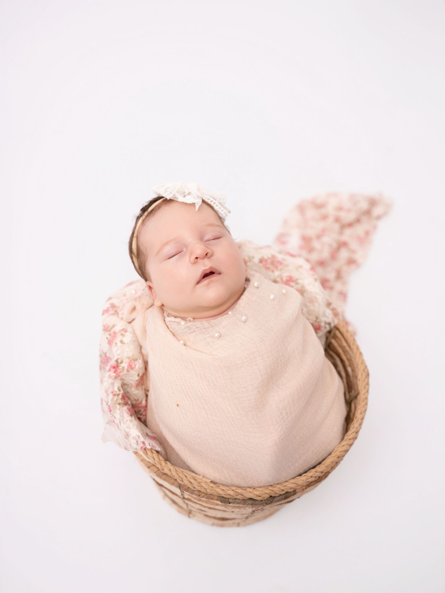 Sleeping newborn baby girl wrapped in a pink floral swaddle with a bow headband, nestled in a wicker basket.
