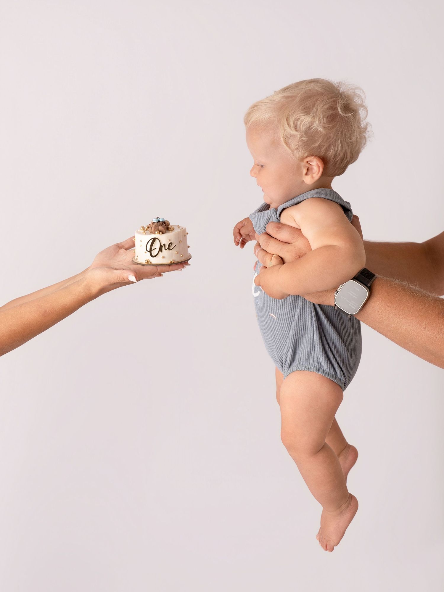 A baby in a gray romper reaches for a smash cake reading One, held by an adult, celebrating their first birthday.