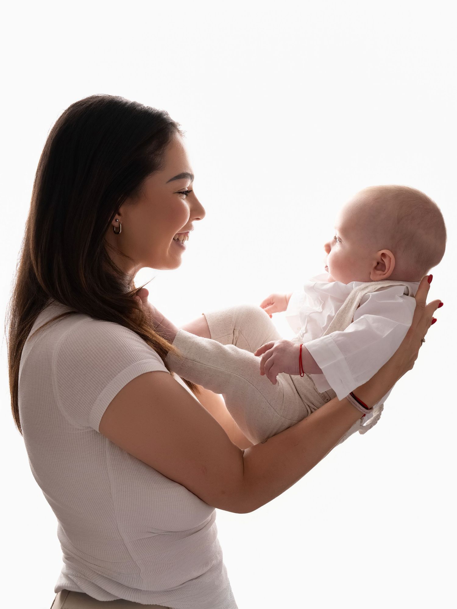 Smiling young mother holding her newborn baby up in a bright white studio setting, side profile view.