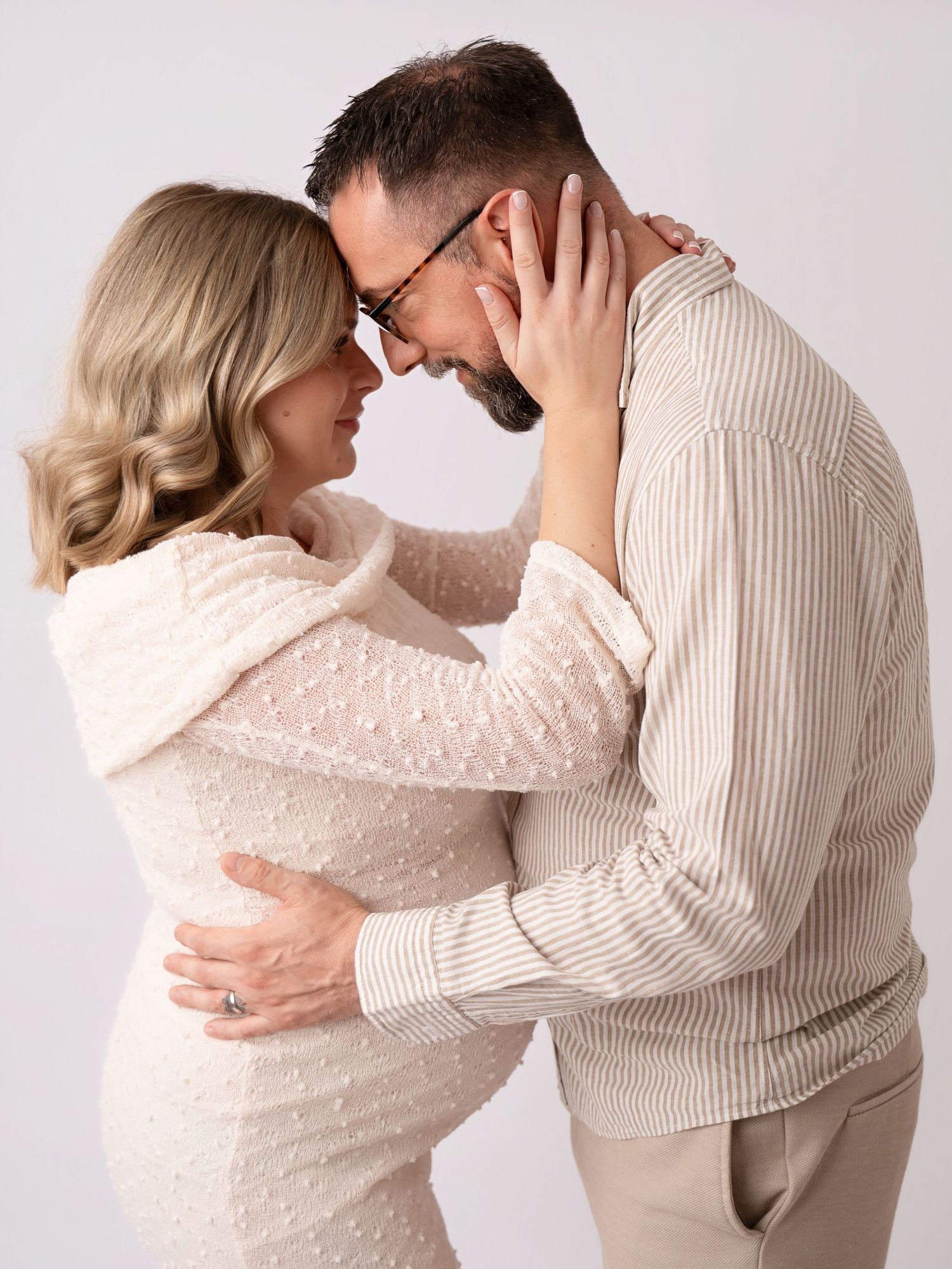 A couple in neutral tones shares a tender forehead-to-forehead moment, embracing in a soft studio setting.
