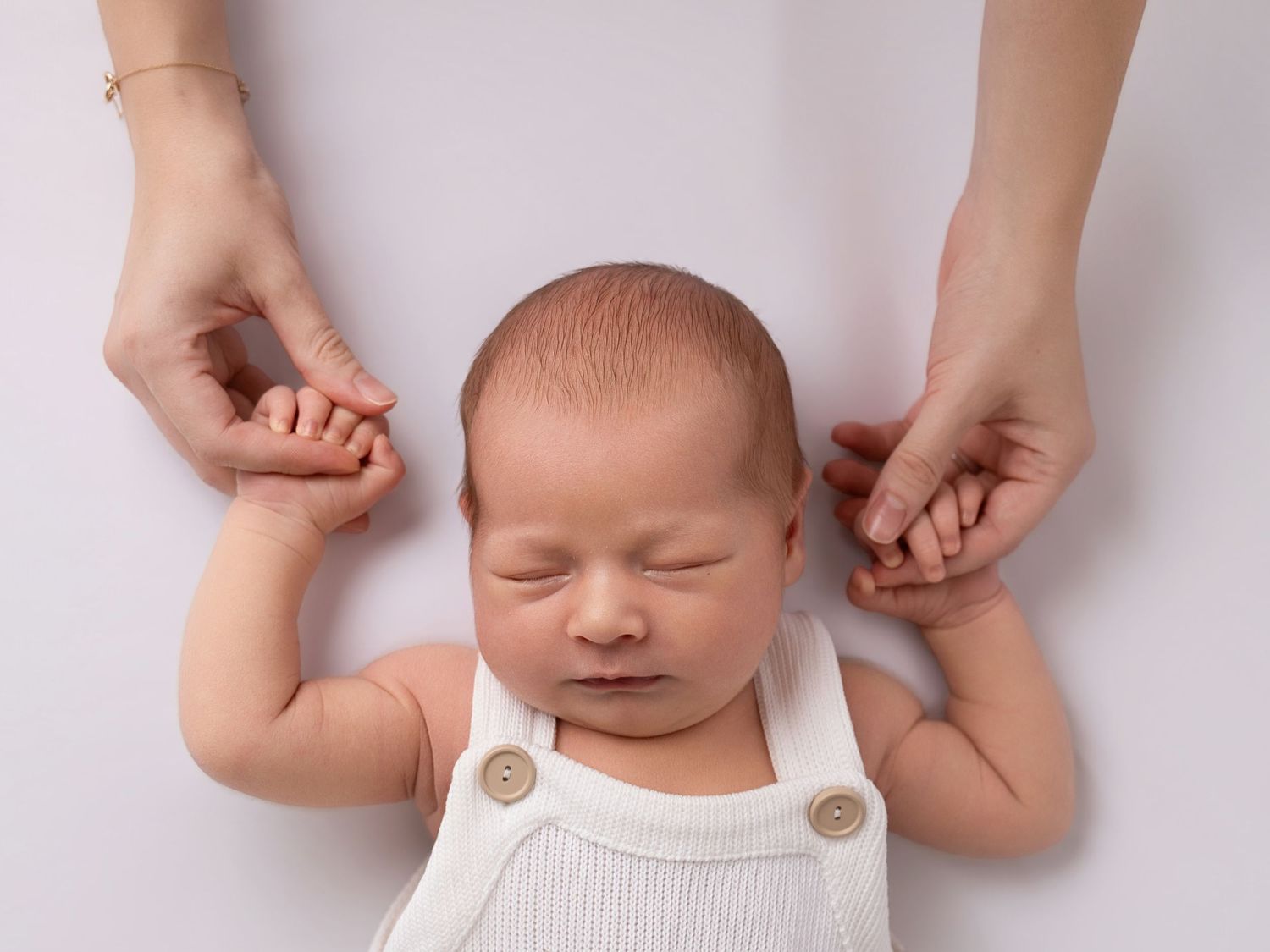 Newborn baby in white romper sleeping peacefully while parent gently holds tiny hands in professional photo.