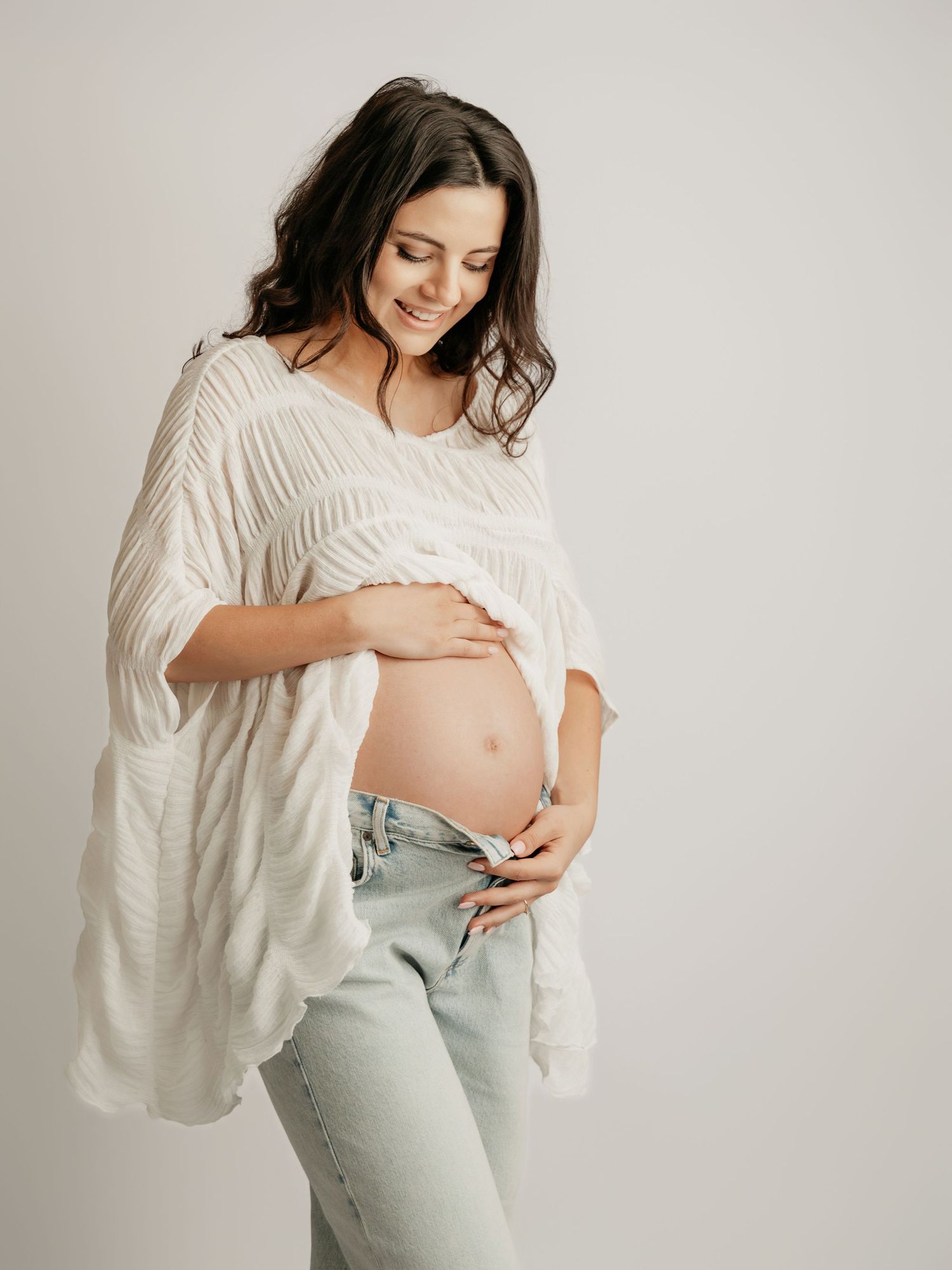 Glowing pregnant woman in white textured top and jeans smiles while holding baby bump in studio.