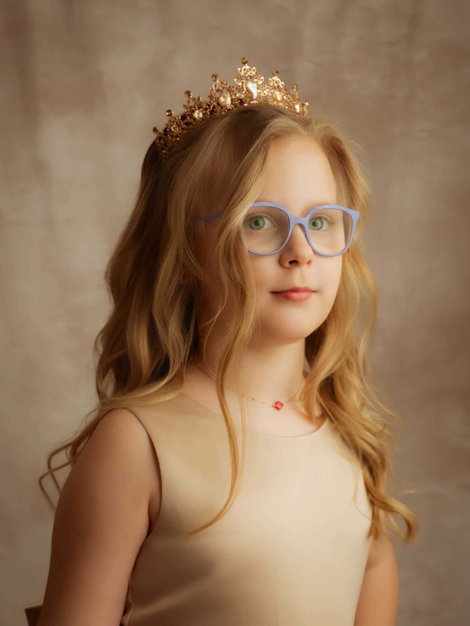 A young girl with wavy blonde hair wearing a gold crown and blue glasses in a studio portrait.