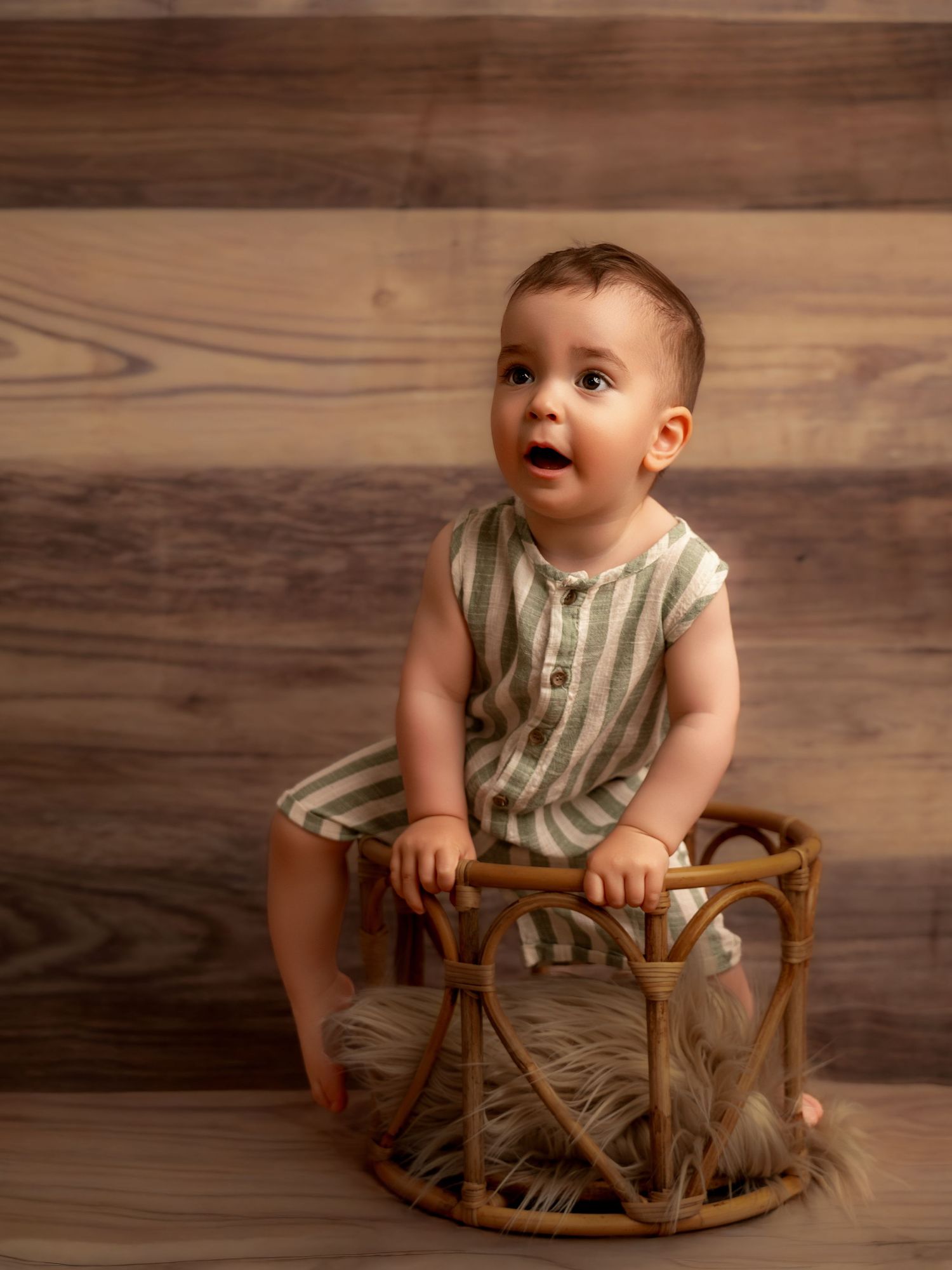 Adorable baby in striped romper sitting in a wicker basket against a rustic wood plank backdrop.