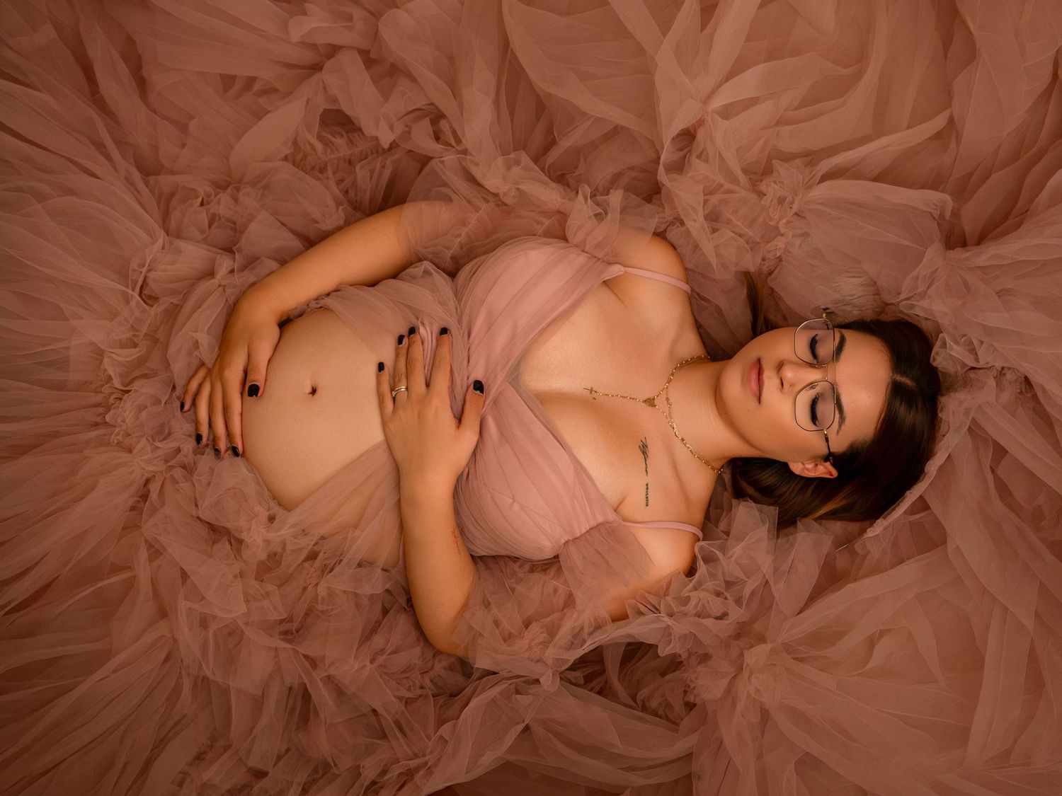 Pregnant woman in pink tulle gown lying down with eyes closed, cradling baby bump in dreamy maternity photo.