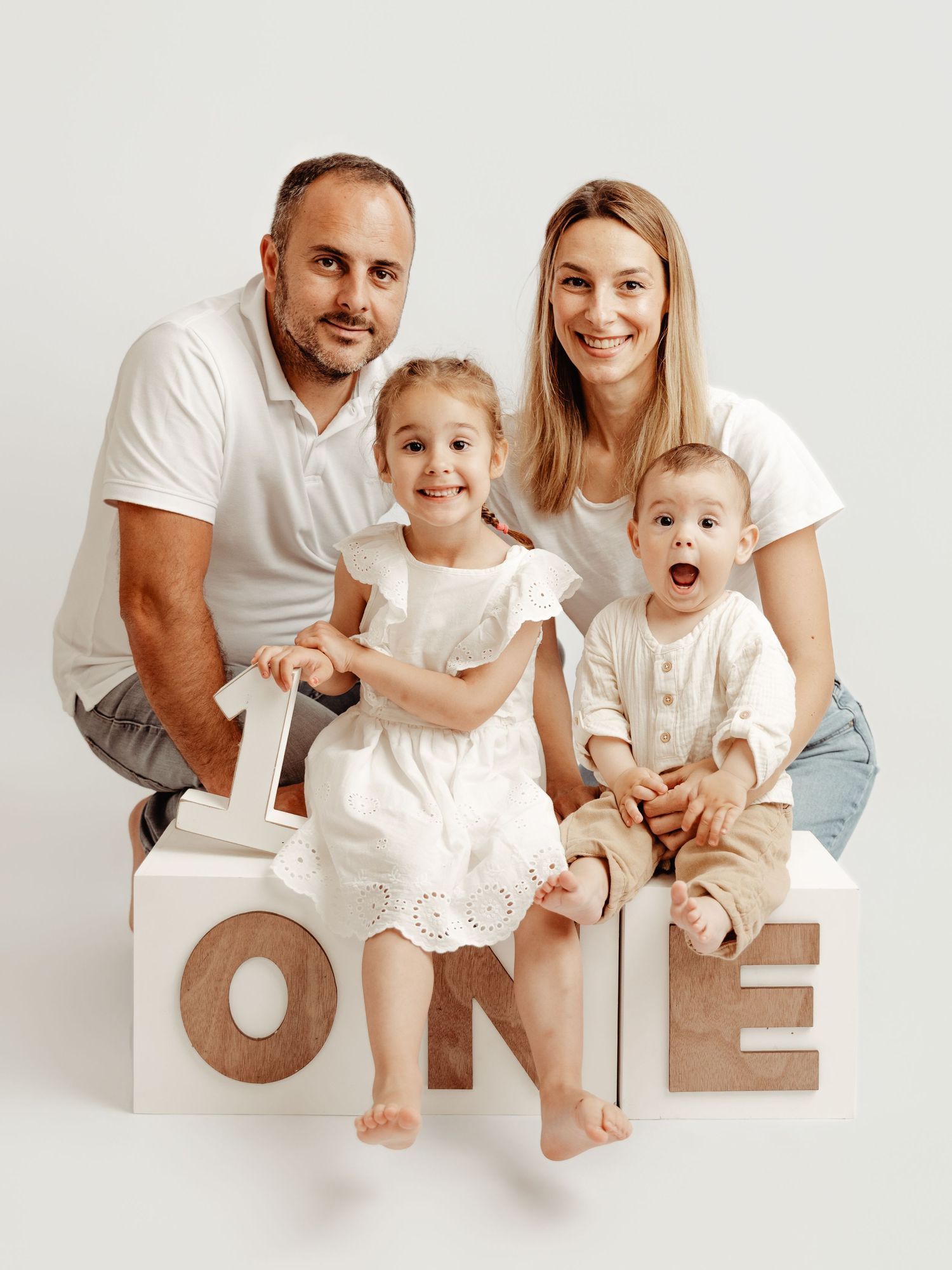 Happy family of four posing with ONE letter blocks, celebrating baby's first birthday in matching white outfits.