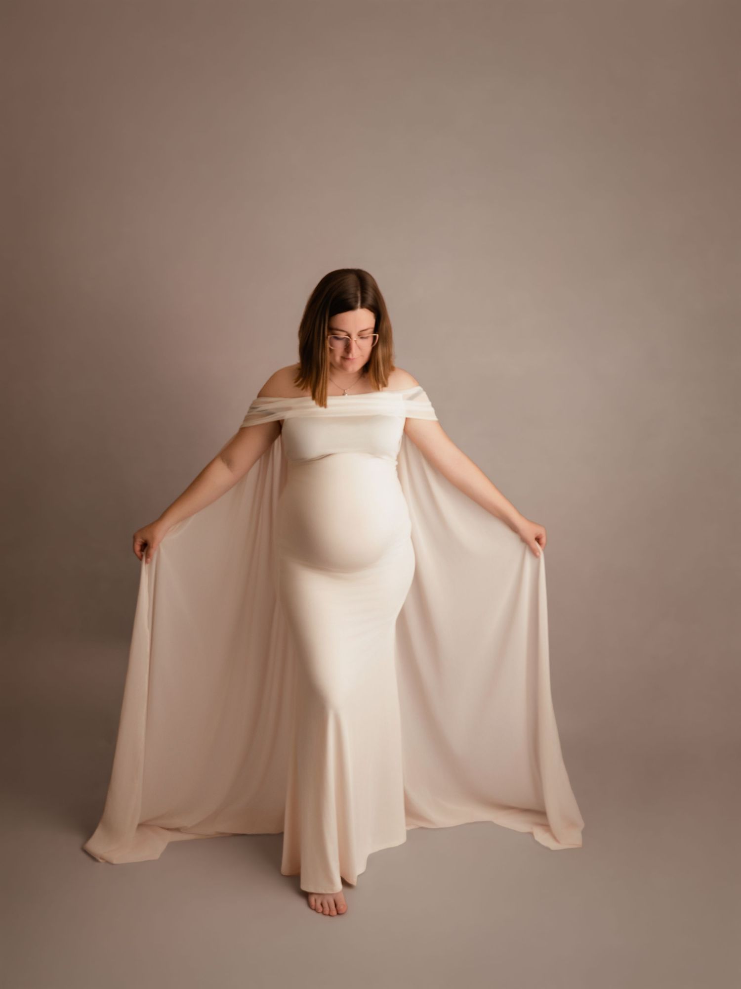 Pregnant woman in elegant off-shoulder white gown with flowing cape sleeves poses gracefully in a studio maternity photo.
