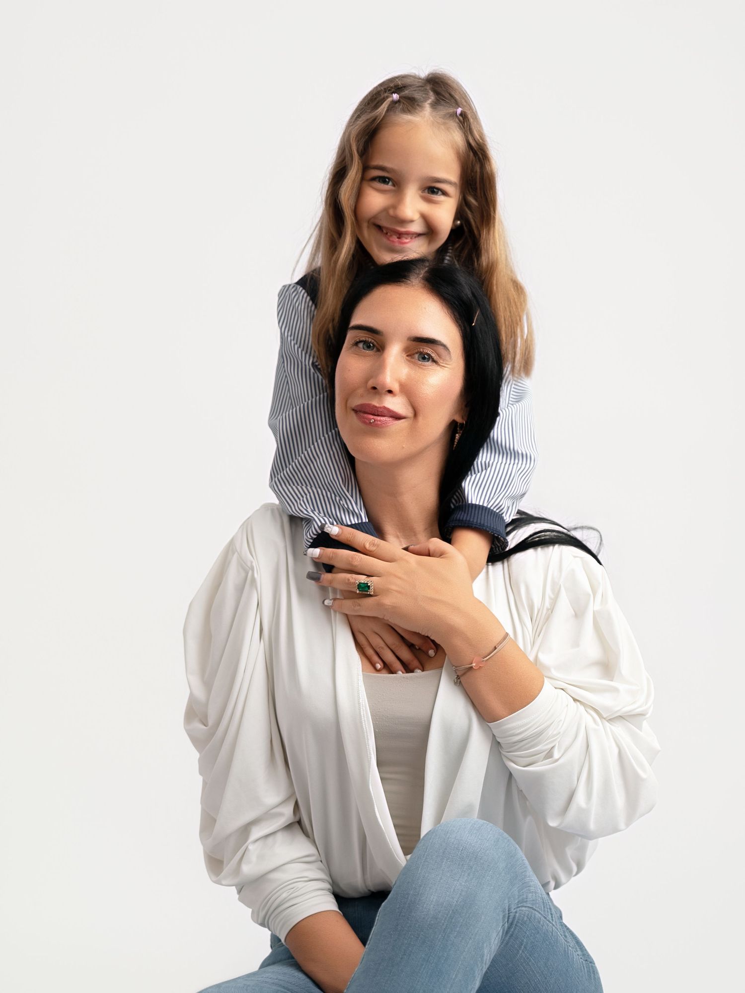 A woman sits smiling with a young girl perched on her shoulders, both posing against a white background.
