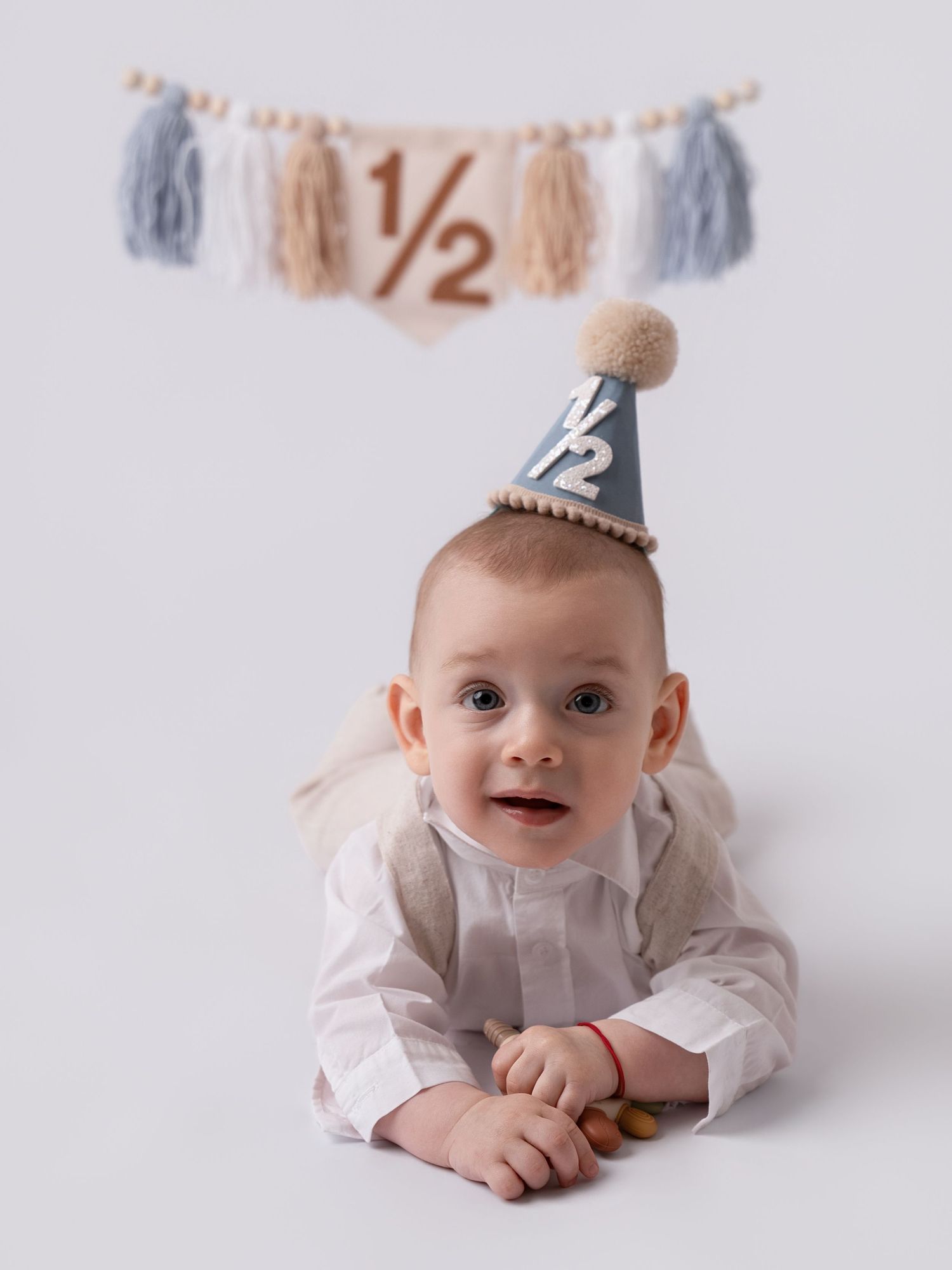 Baby in a blue half-birthday party hat lying on tummy, with a decorative 1/2 banner and tassels in the background.