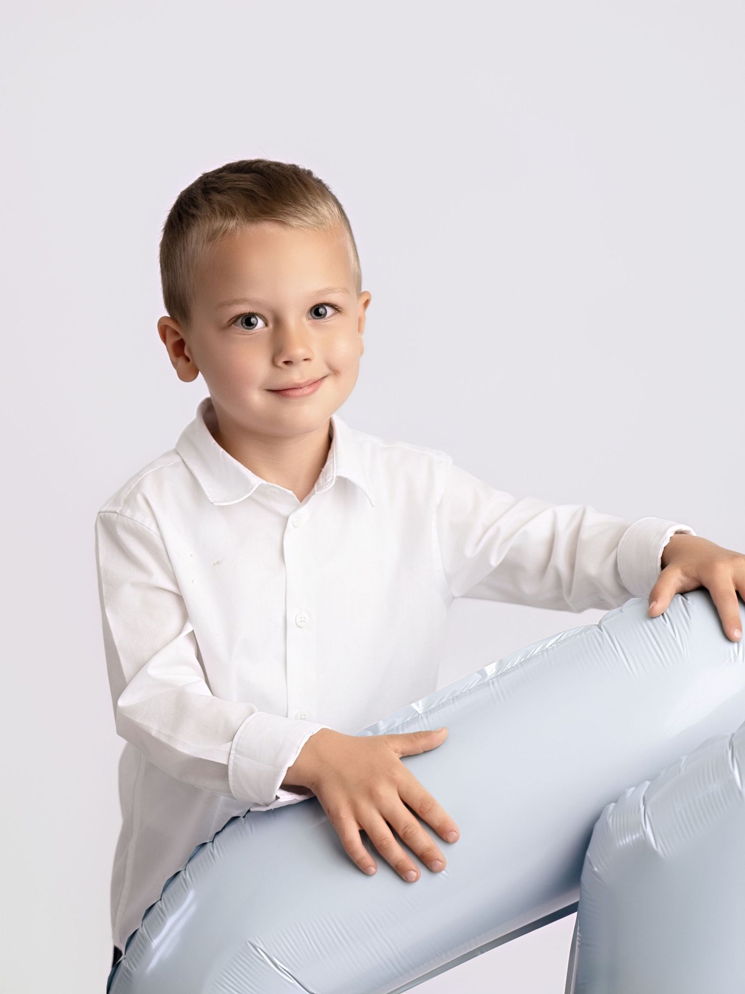 Young boy in white shirt smiling while leaning on a light blue number balloon against white background.