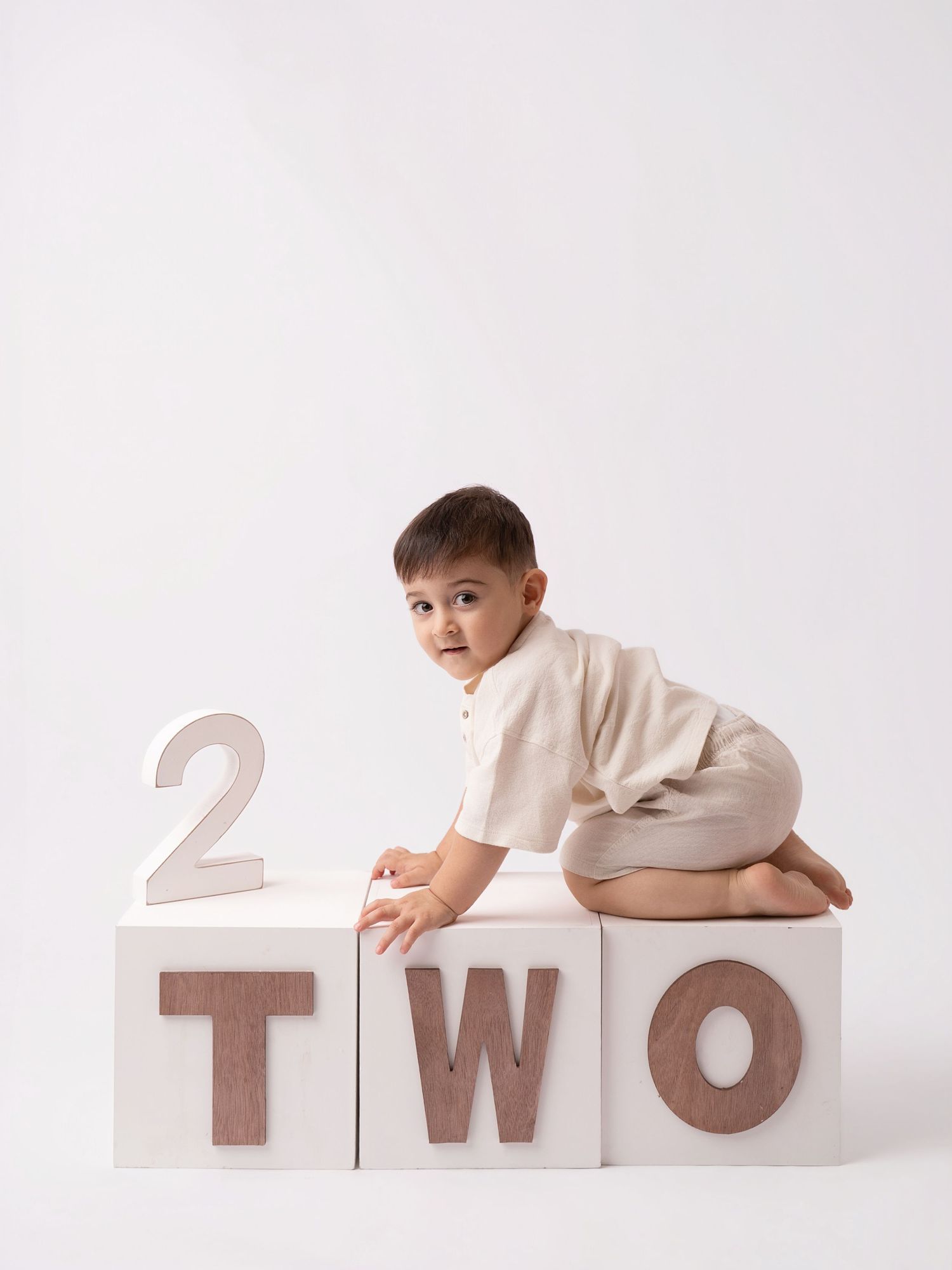 A toddler in a beige outfit sits on white blocks spelling TWO, celebrating their 2nd birthday.