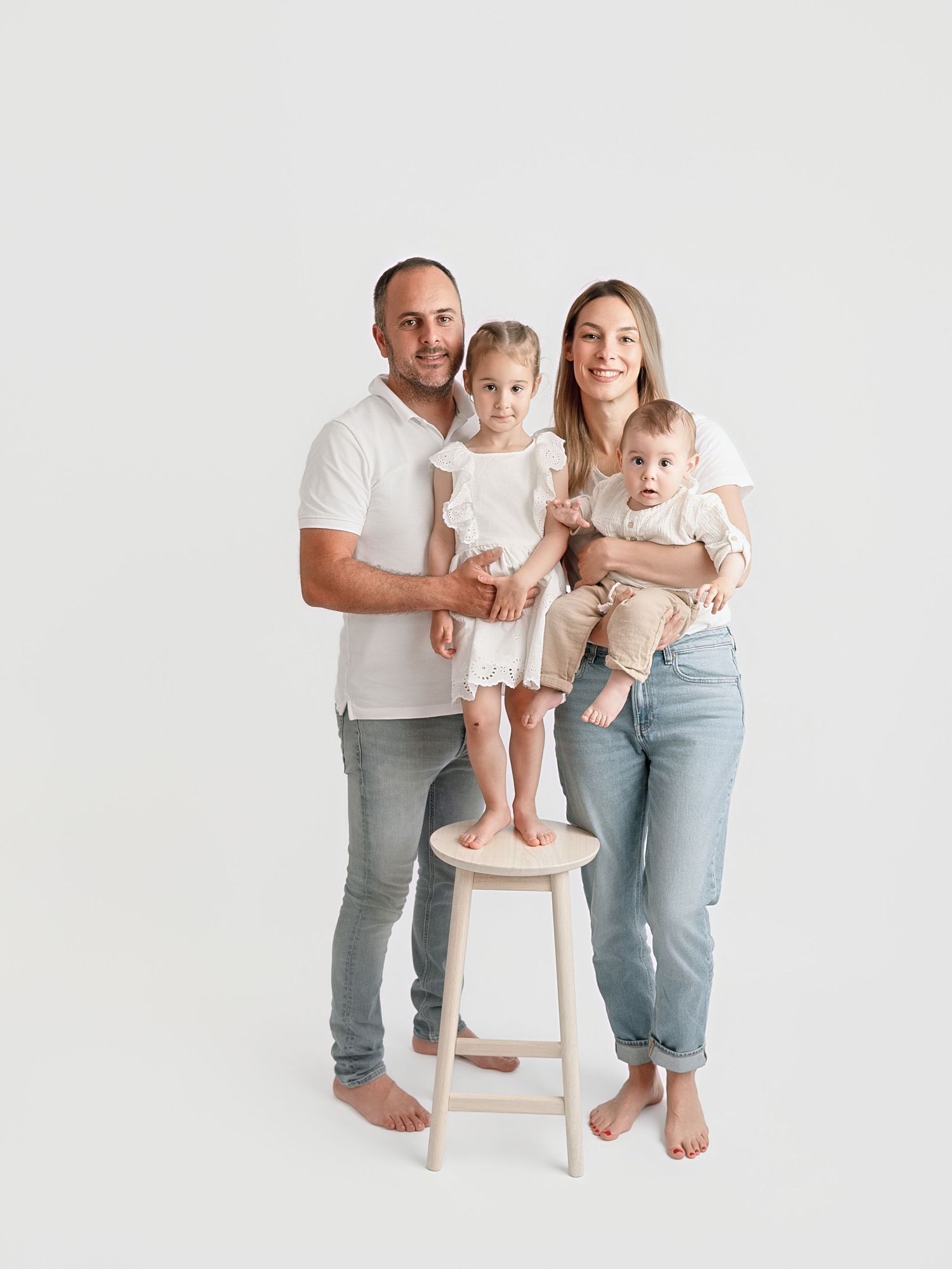 Family of four posing in white studio, parents in white tops and jeans with toddler on stool and baby in arms.